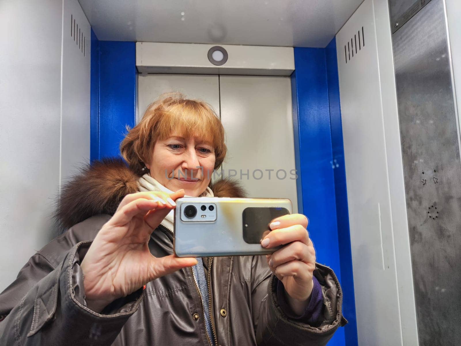 Middle-aged woman in warm jacket and a hood with fur taking selfie in mirror in elevator. Female Traveler in Alaska, Lapland, Arctic, Siberia