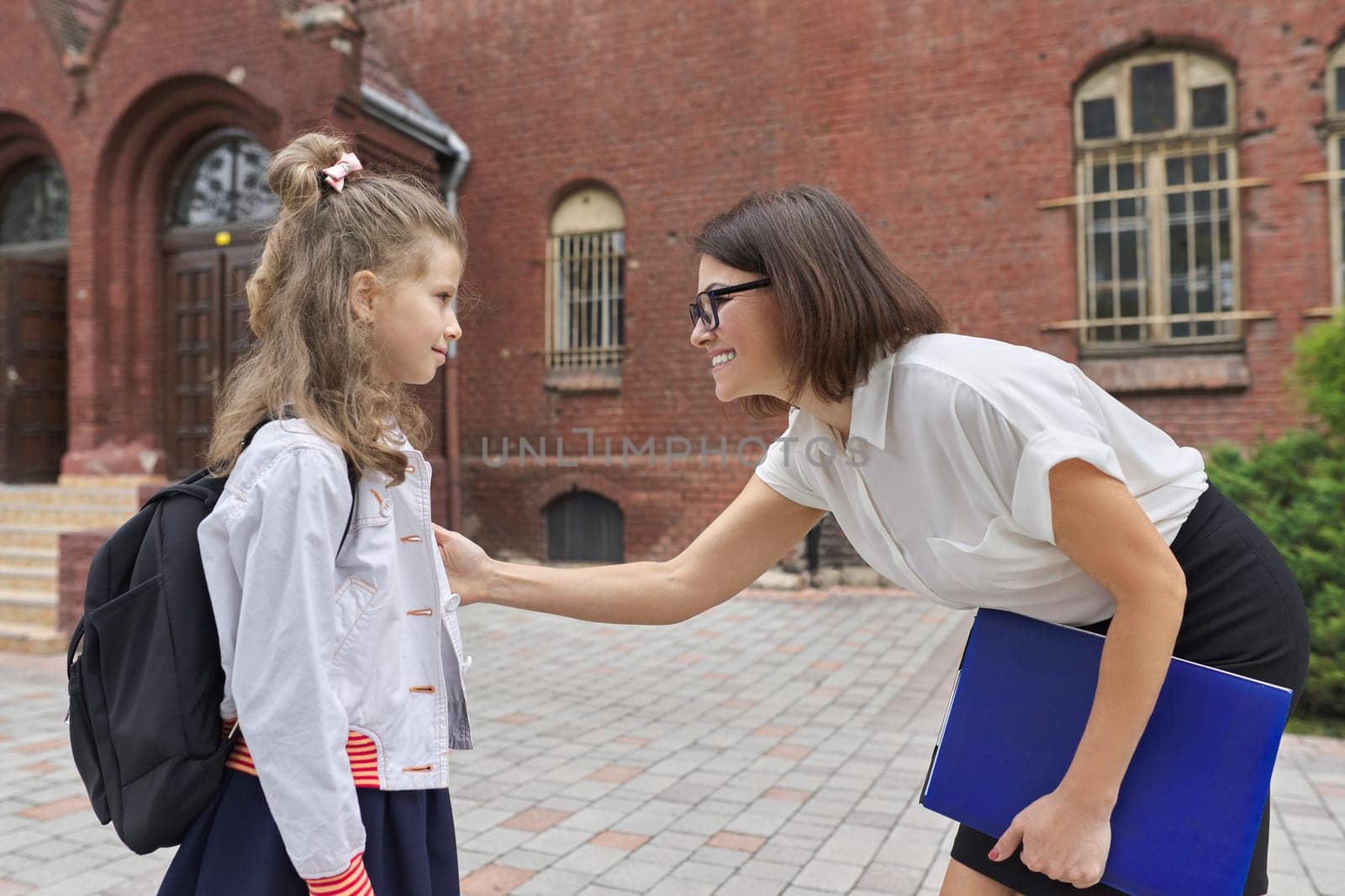 Outdoor portrait of teacher woman and little student girl together. Teacher talking with child near school building. Back to school, start of classes