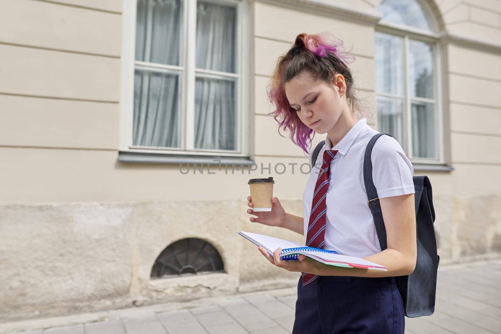 Girl student 15, 16 years old walking with backpack, reading notebook and drinking drink. Back to school, college, y spacecop