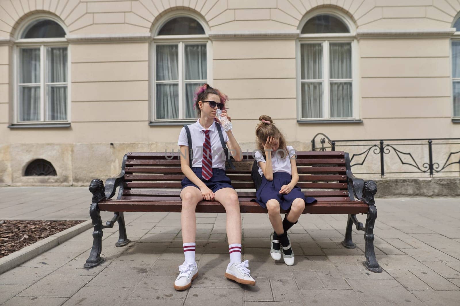 Tired schoolchildren sitting on bench. Two girls sisters teenager and elementary school student resting after class at school. Fatigue, stress, problems in school life