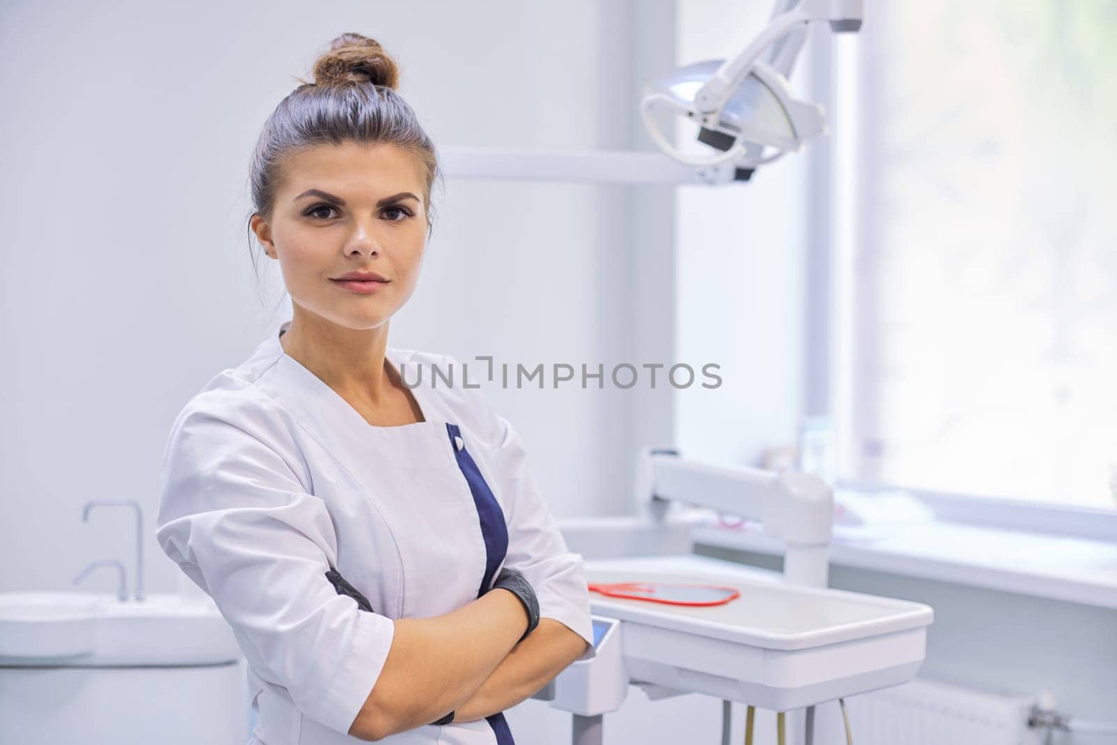 Young confident female dentist doctor with folded arms, dentistry cabinet background, copy space