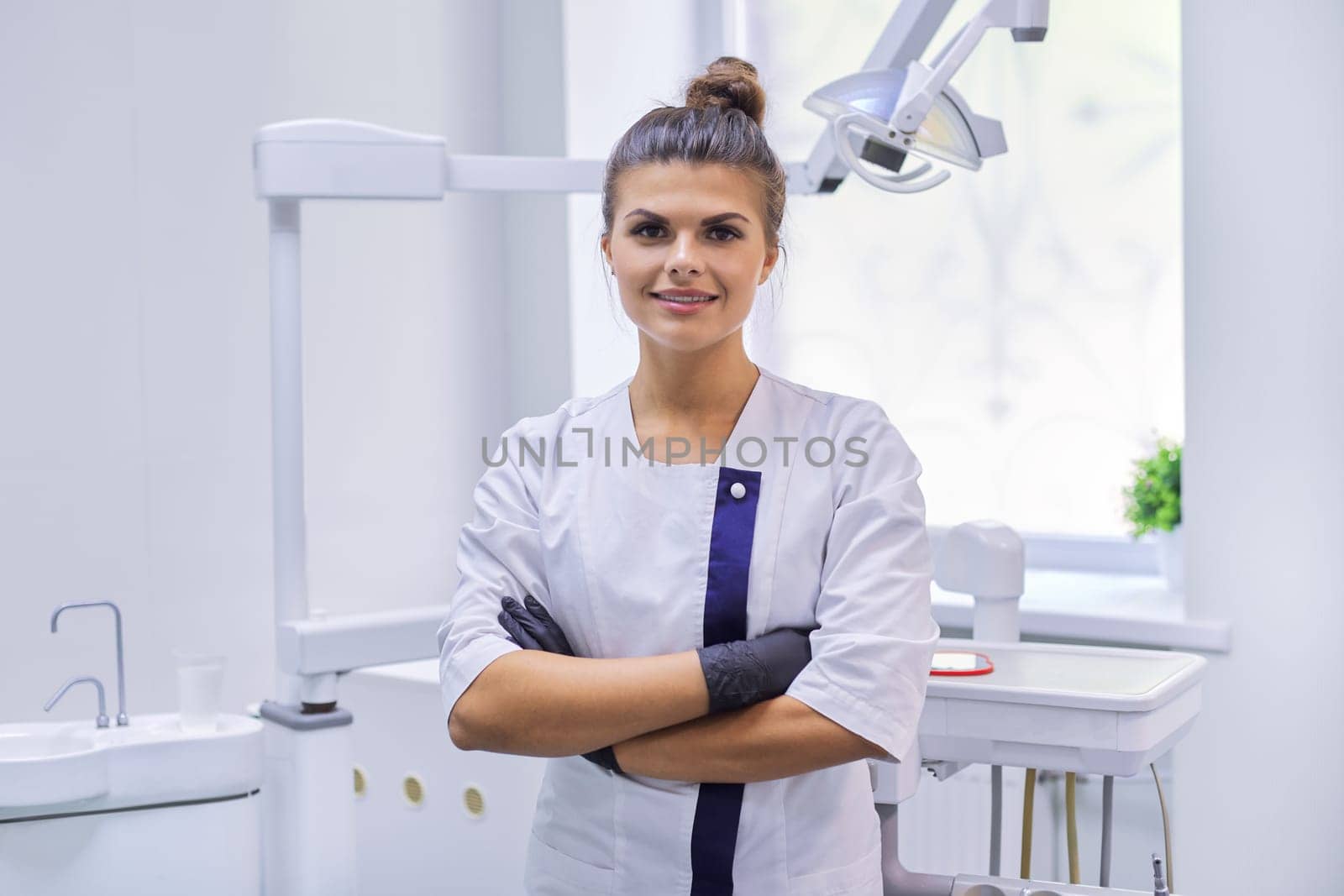 Portrait of young confident smiling dentist doctor woman, female with arms crossed looking at camera standing in dentistry office