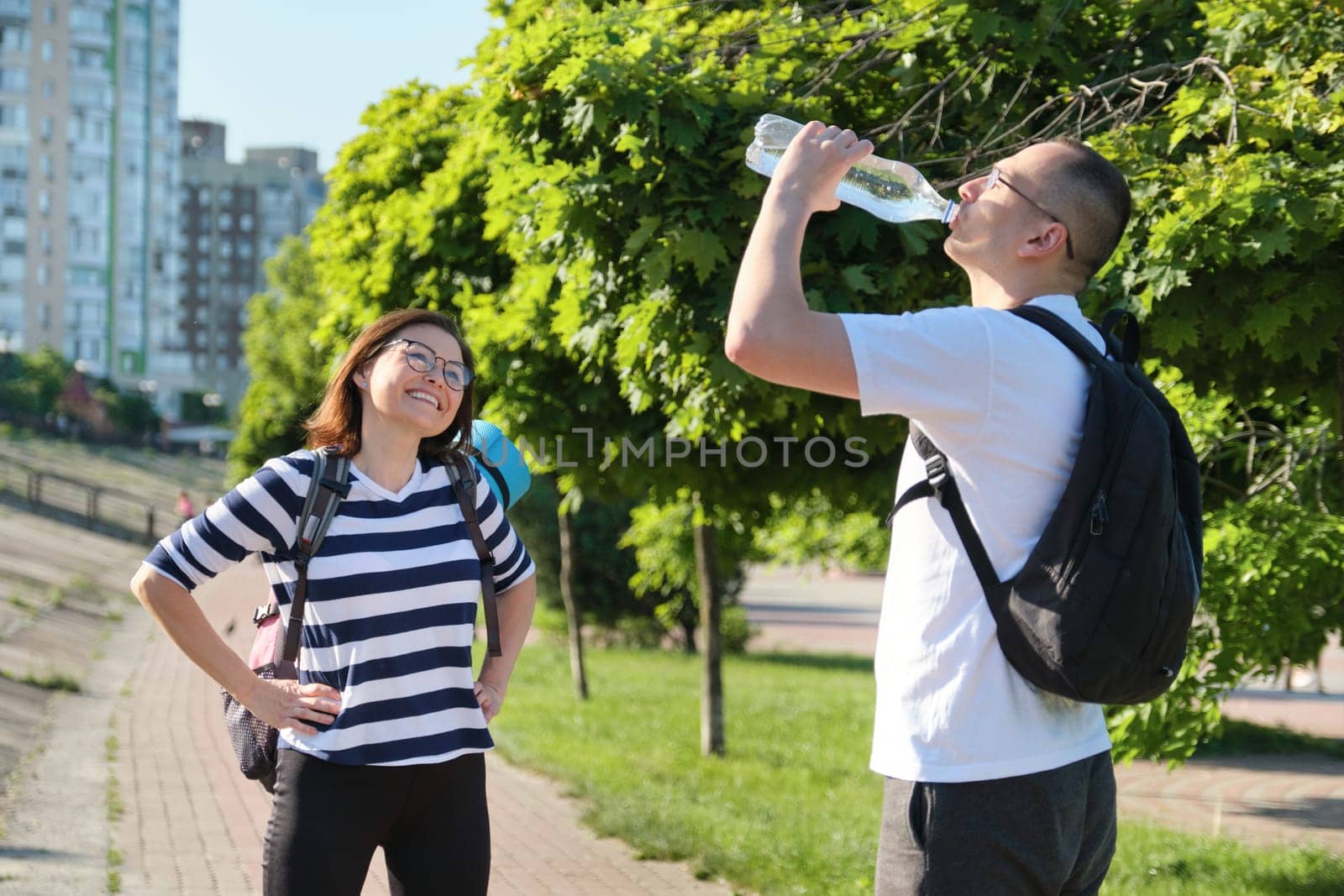 Mature smiling man and woman in sportswear with backpacks exercise mat walking in city park talking drinking water from bottle, active healthy lifestyle of middle-aged people