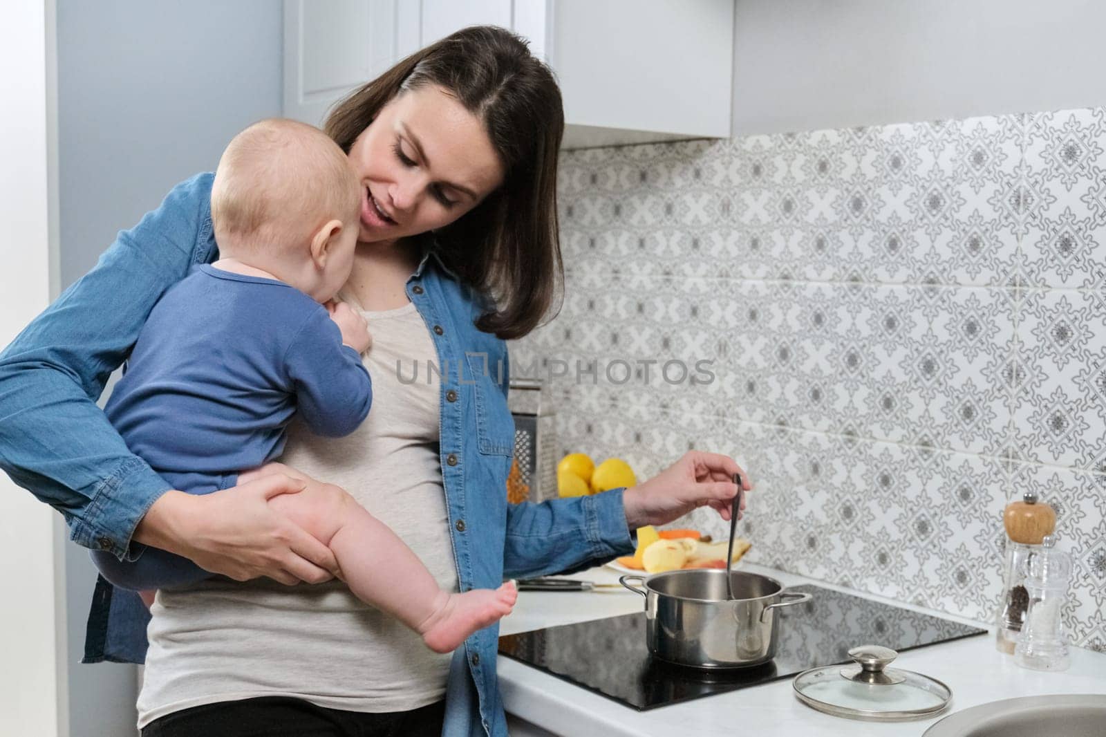 Young beautiful woman with baby in her arms in kitchen. Mother with son cooking food in pan in home kitchen, healthy home food and parent child communication