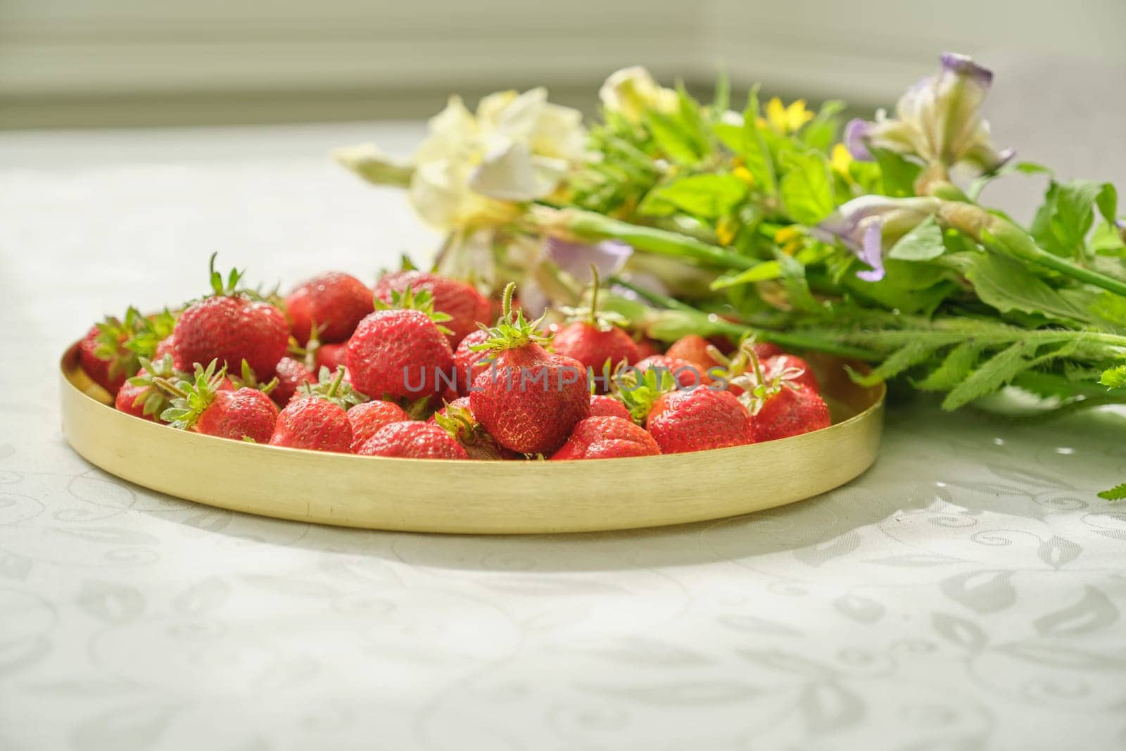 Berries of red ripe strawberry on white table in golden tray, natural vitamins in spring season, healthy eating. Bouquet of flowers lying near strawberries