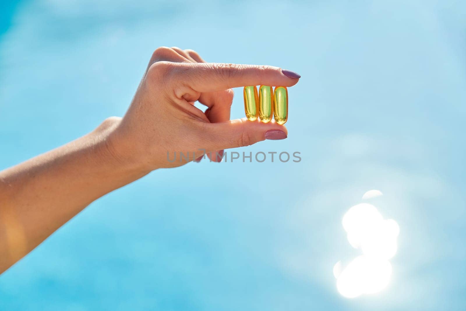 Sunny golden vitamin D oil capsules Omega-3 in woman hand, background sun blue water. Healthy lifestyle, nutritional supplements, diet
