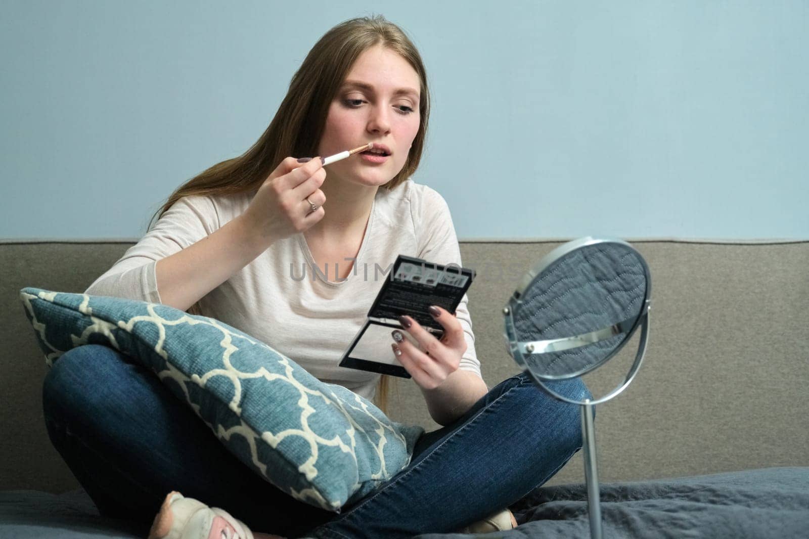 Young beautiful blonde woman doing makeup with make-up mirror and brush, sitting at home on bed, copy space