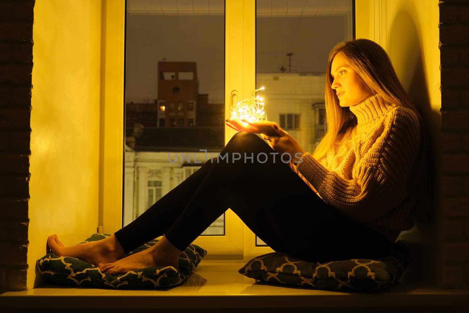 Winter portrait of young beautiful woman sitting near window on windowsill, night city background, sparkling garland lights in hands of girl