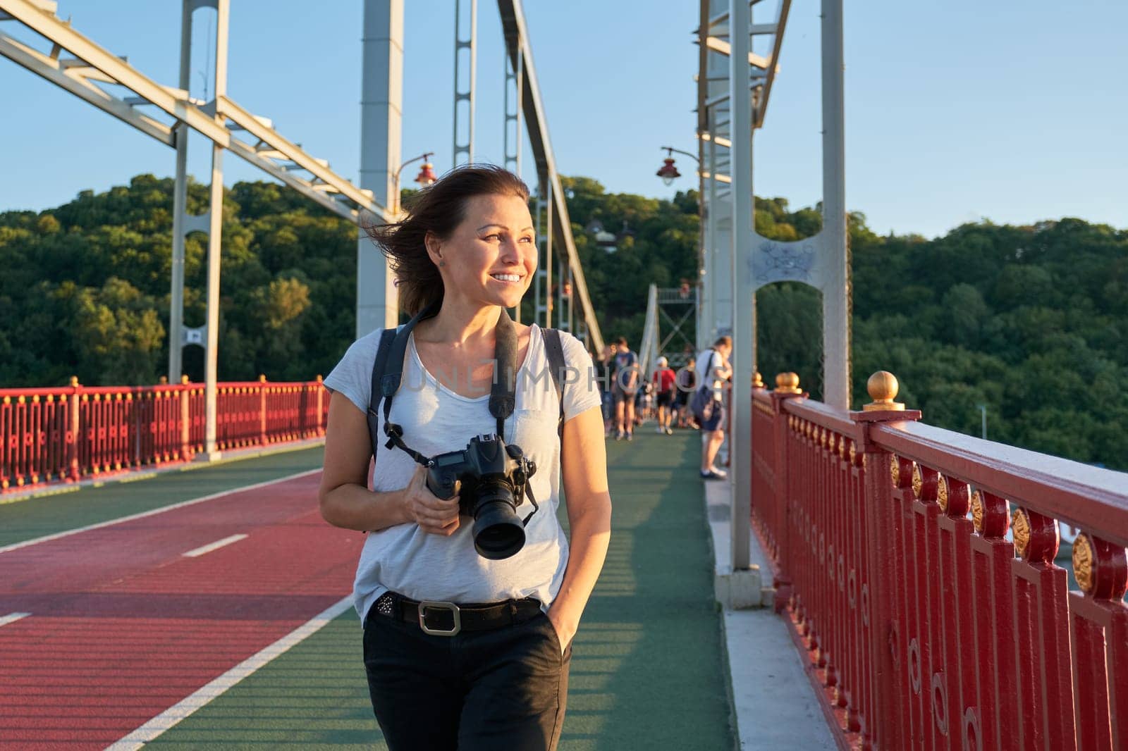Positive middle-aged woman walking on bridge. Mature female with backpack photo camera smiling on sunny summer day