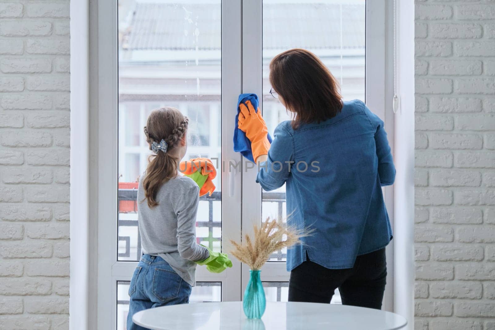 Mother and daughter child in rubber gloves with detergent and rag washing windows together. Girl helping woman do house cleaning