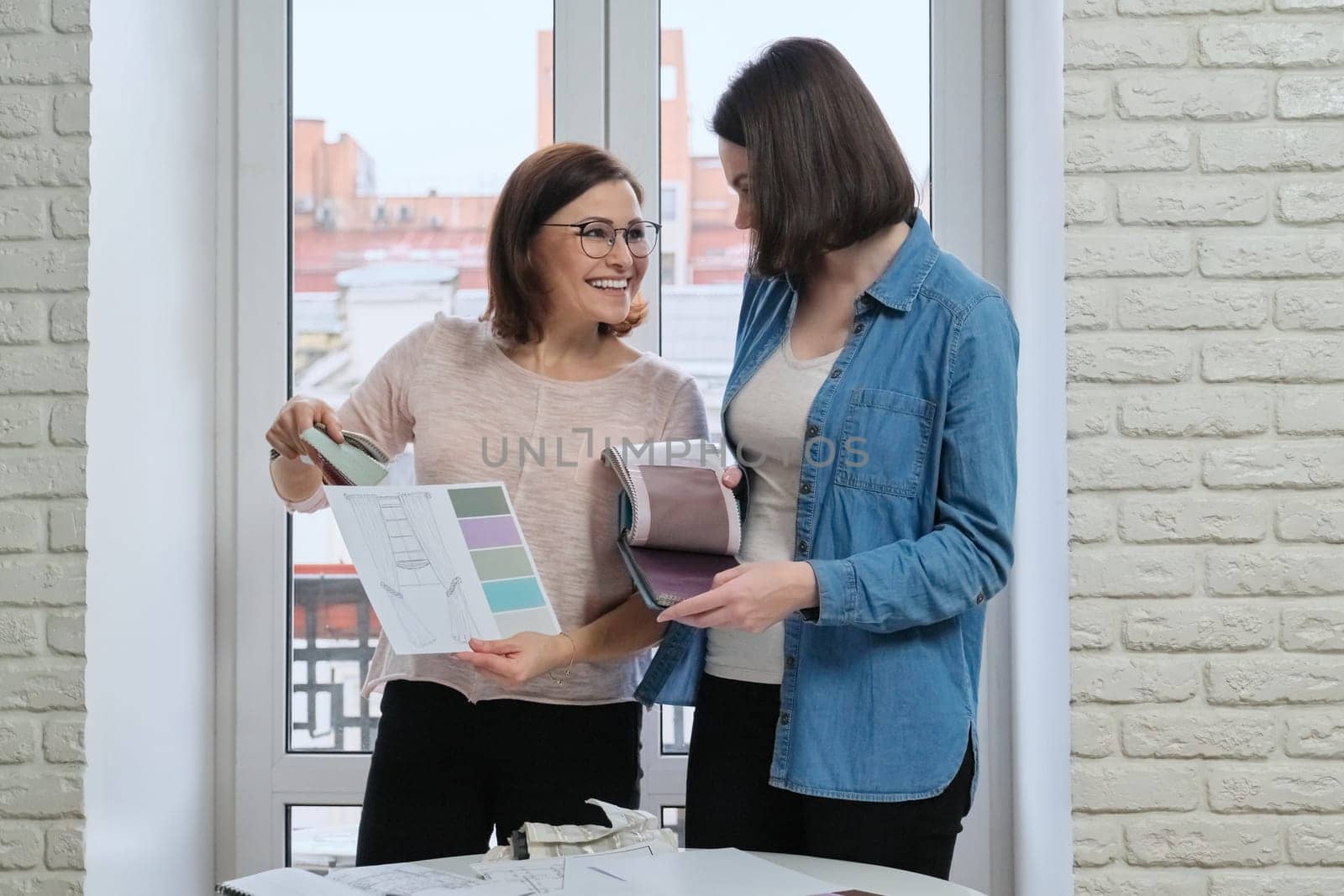 Female designer and client working with fabric samples. Selecting fabrics and design of curtains, females near window with sketch and materials