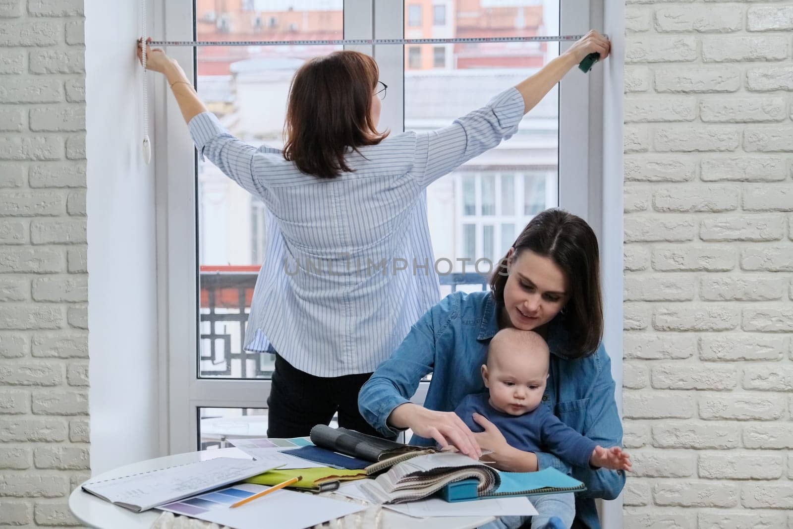 Window decoration with curtains, woman designer taking window sizes. At the table, sitting mother and baby choosing fabrics, color, model