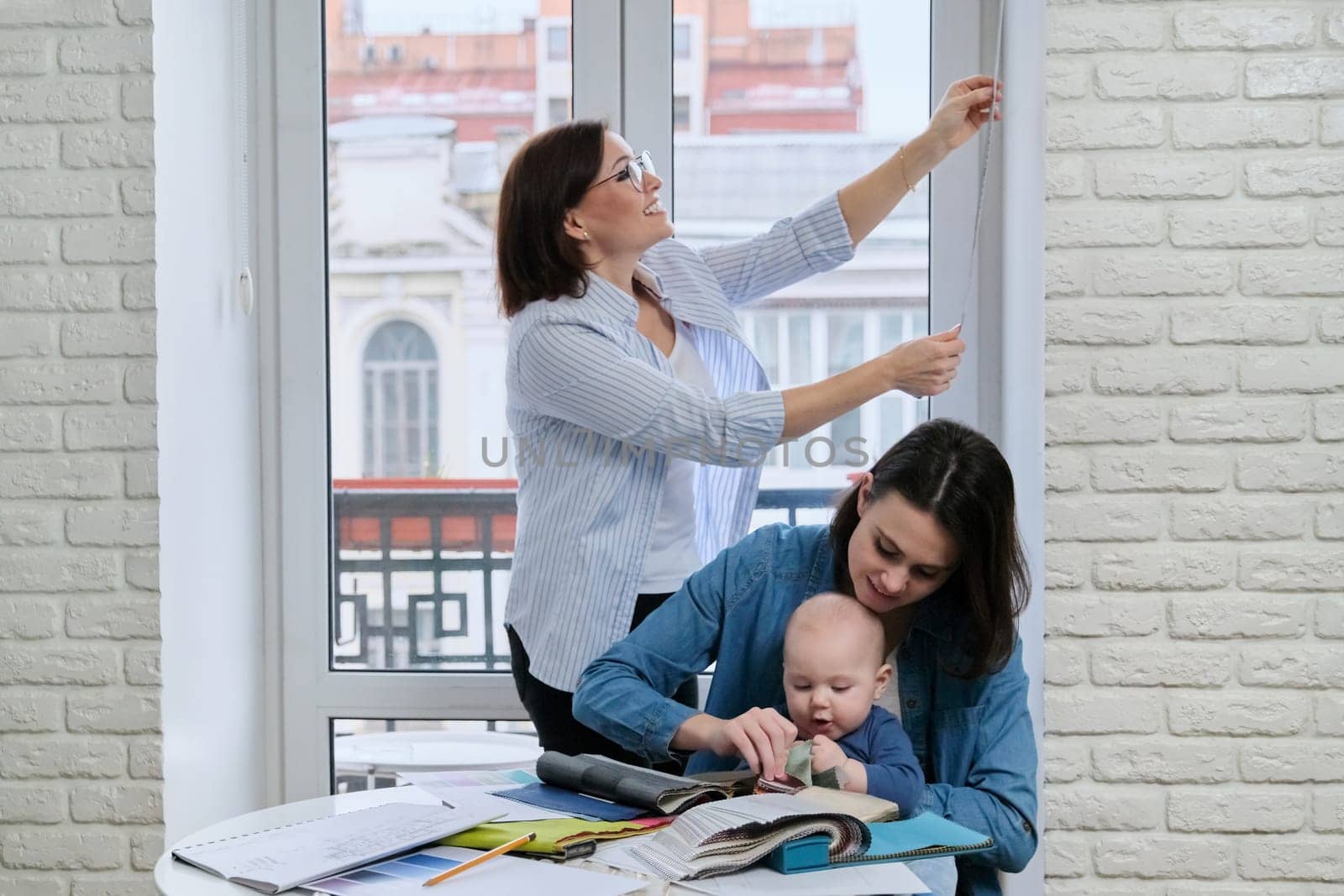 Window decoration with curtains, woman designer taking window sizes. At the table, sitting mother and baby choosing fabrics, color, model