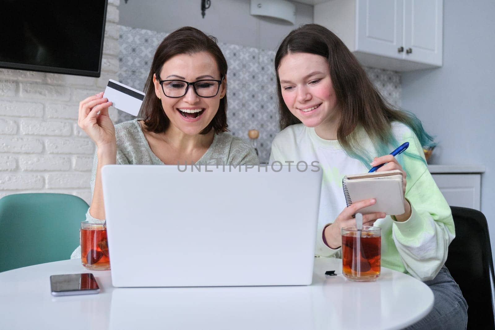 Family, mother and teenage daughter using laptop computer credit card and doing online shopping. Women sitting at home in kitchen talking smiling choosing gifts
