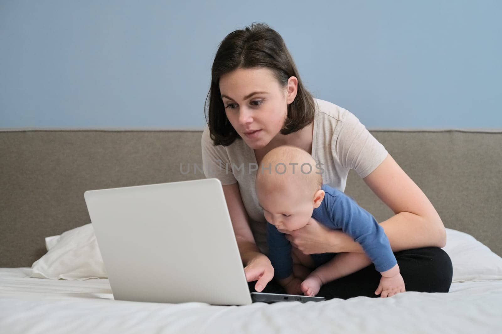 Young mother with baby looks in laptop computer lying at home in bed by VH-studio