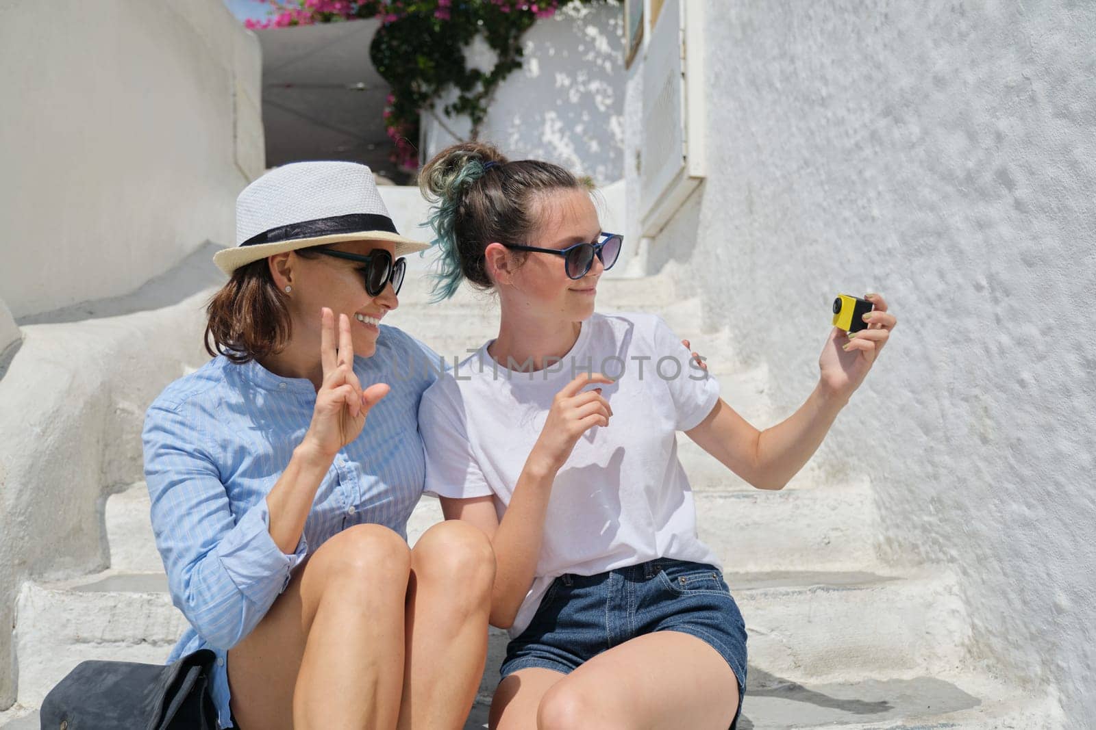 Two women mother and daughter teenager traveling together and recording video vlog on video camera, females sitting on stairs, background white Mediterranean architecture