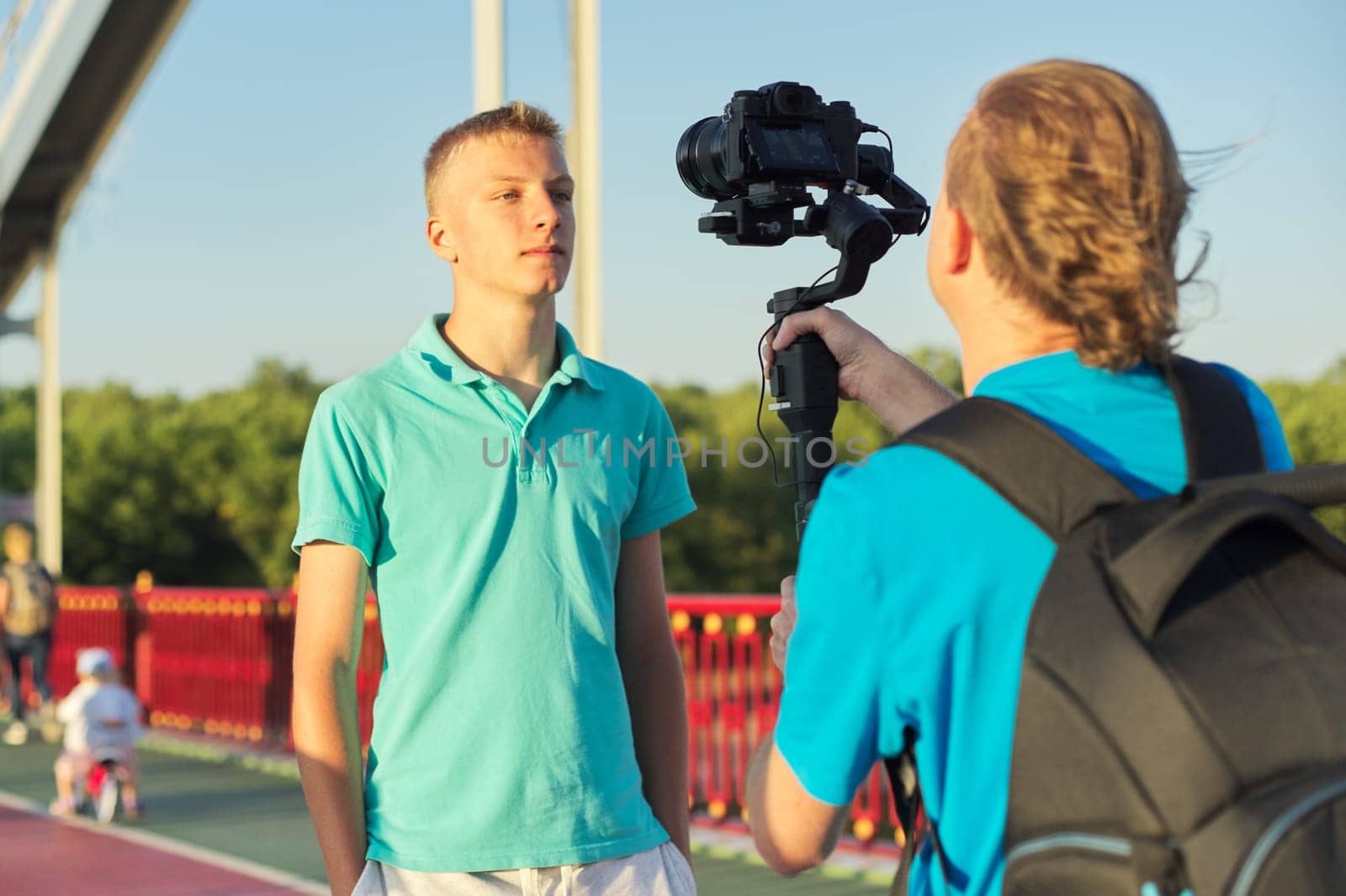 Young man model and male operator taking photos videos with camera and stabilizer, filming backstage, background foot bridge over the river on sunny summer day