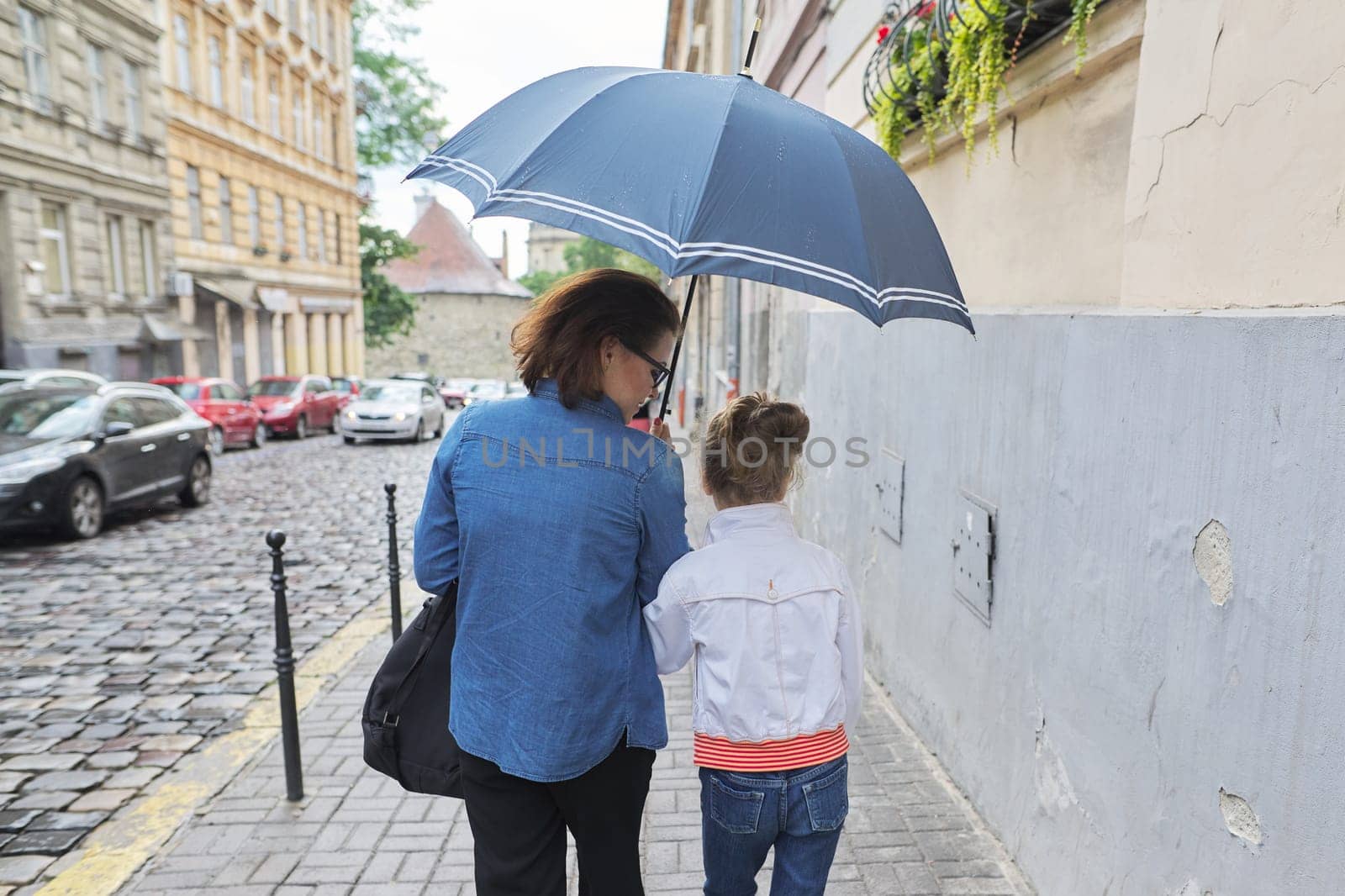 Woman with child girl walking under an umbrella in street by VH-studio