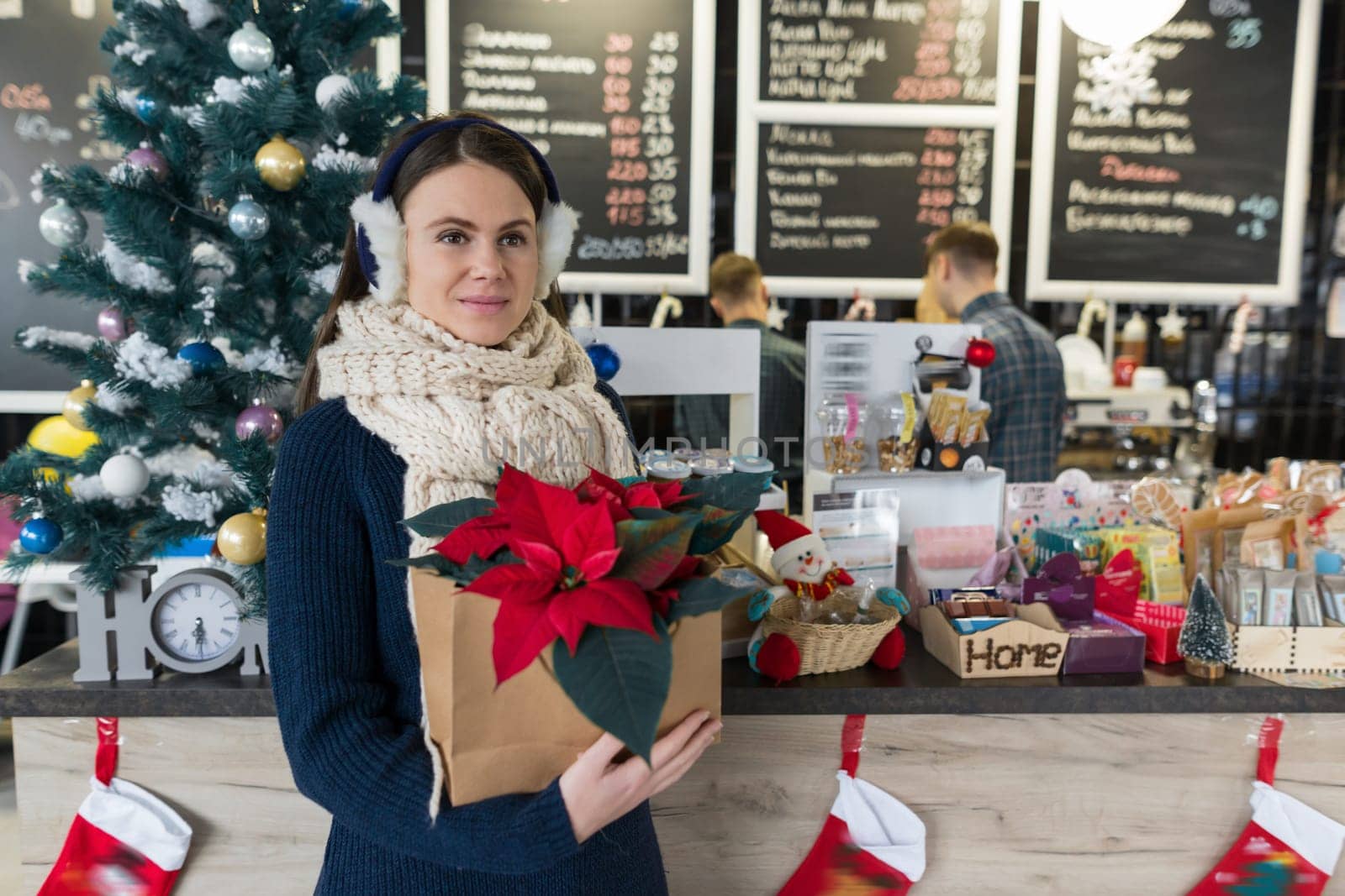 Winter portrait of young beautiful woman in knitted scarf, fur ears, warm sweater with Christmas red poinsettia flower. Girl near bar counter, Christmas decorated tree in coffee shop