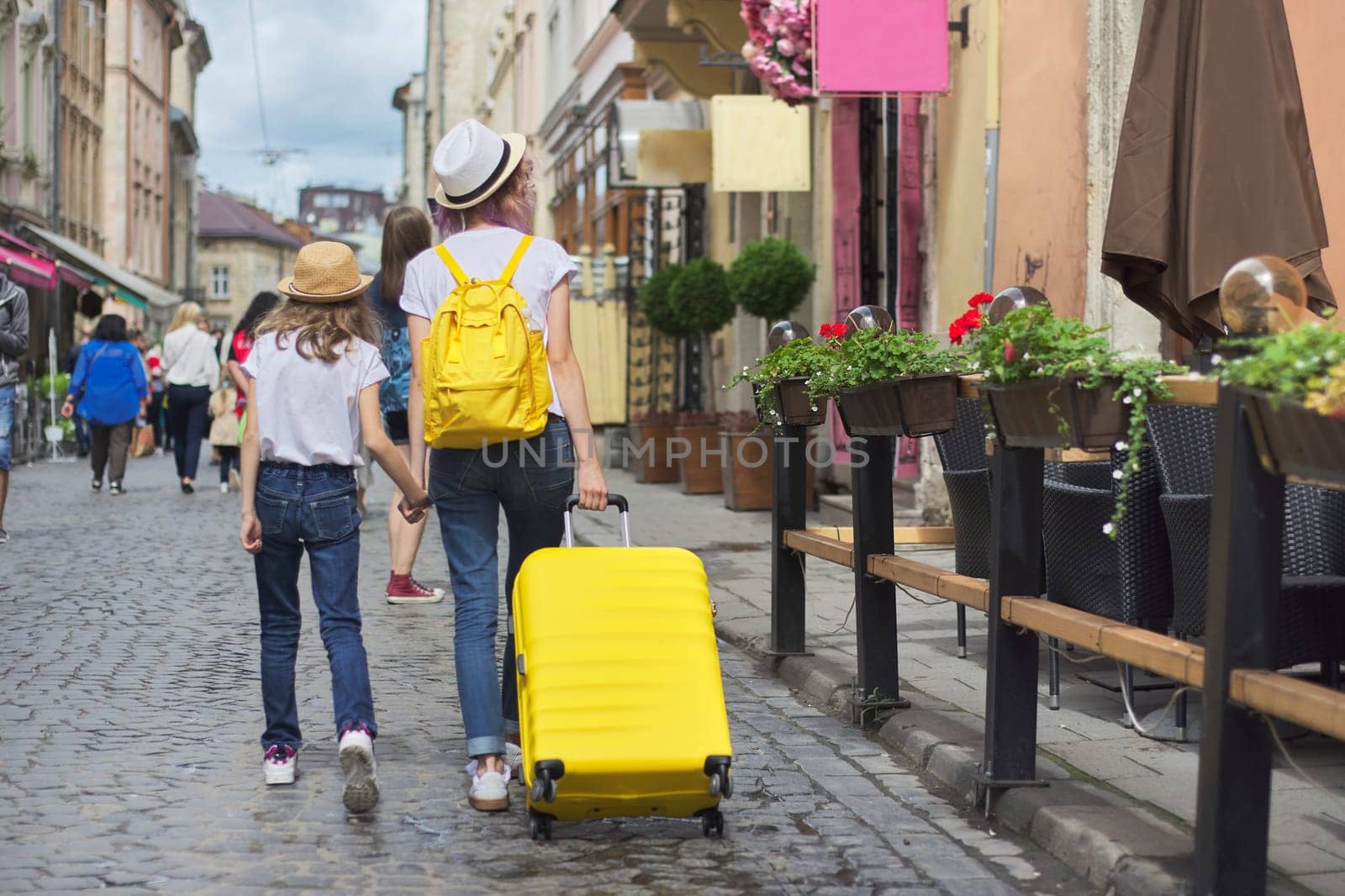 Two girls walking in tourist city holding hands with yellow suitcase, back view