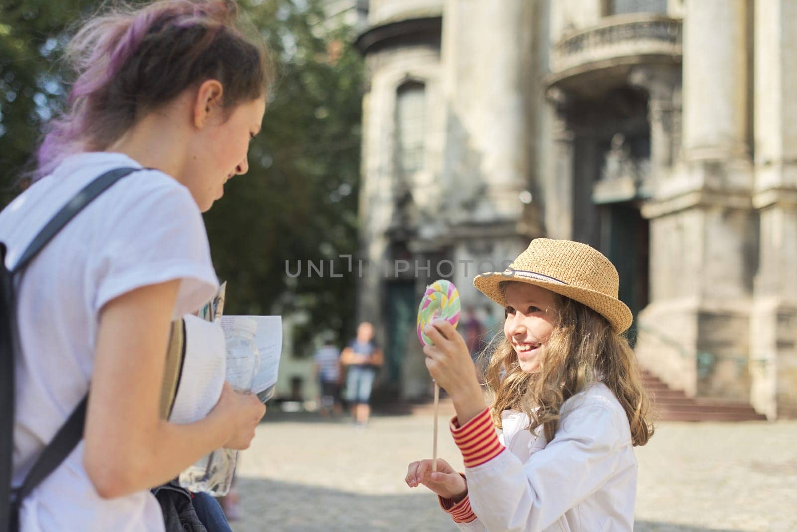 Two laughing girls children sisters on city street, youngest with big colorful lollipop