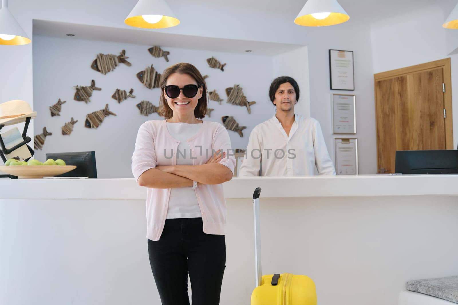 Portrait of female guest with suitcase, male hotel worker near reception, resort hotel interior background