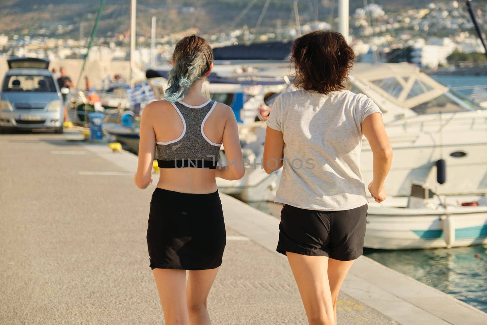 View of the back of mother and daughter jogging running at seaside promenade together, sports outdoor, background sea mountains city, sunny summer day