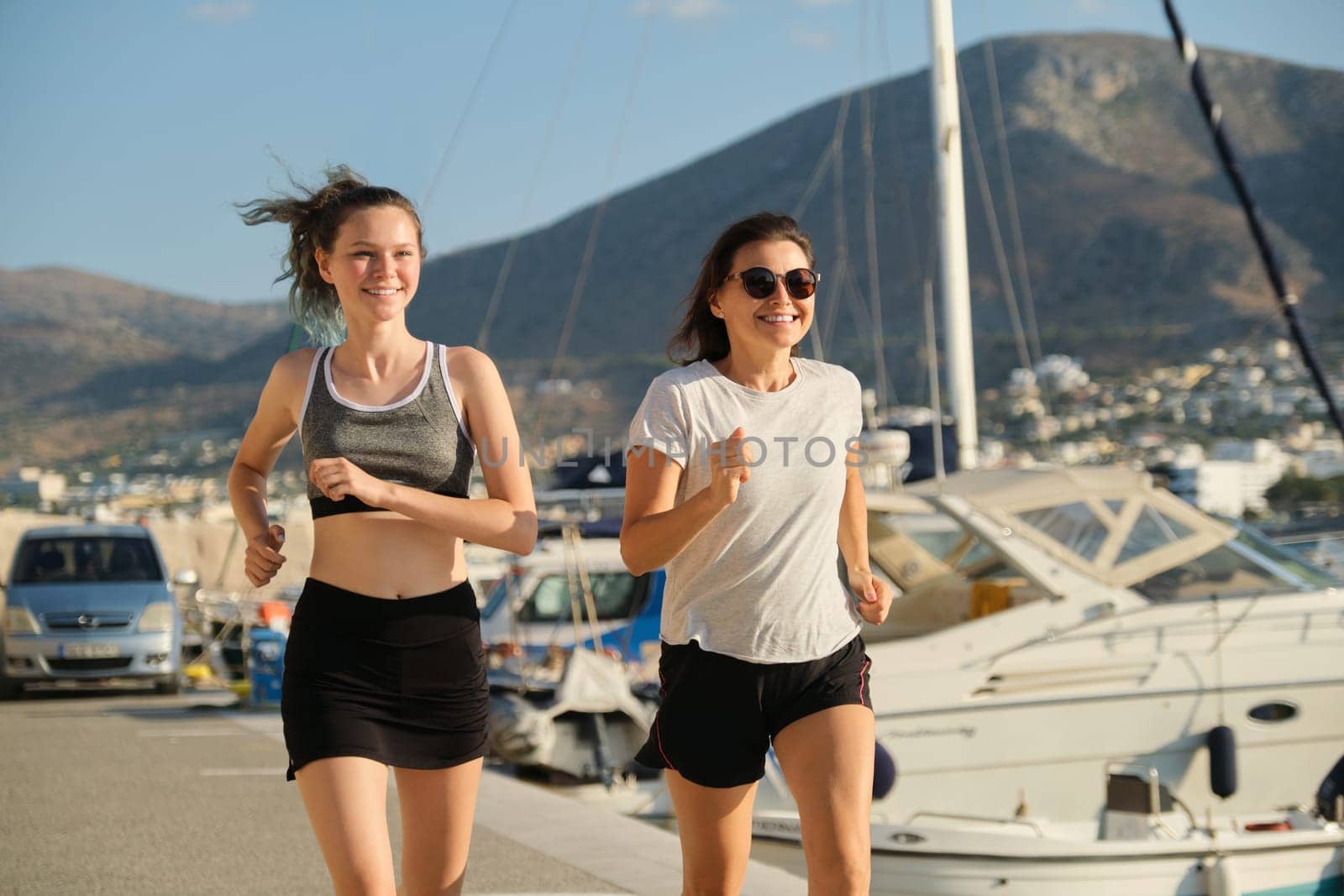 Smiling mother and daughter teenager jogging running at seaside promenade together, sports outdoor, background sea mountains city, sunny summer day