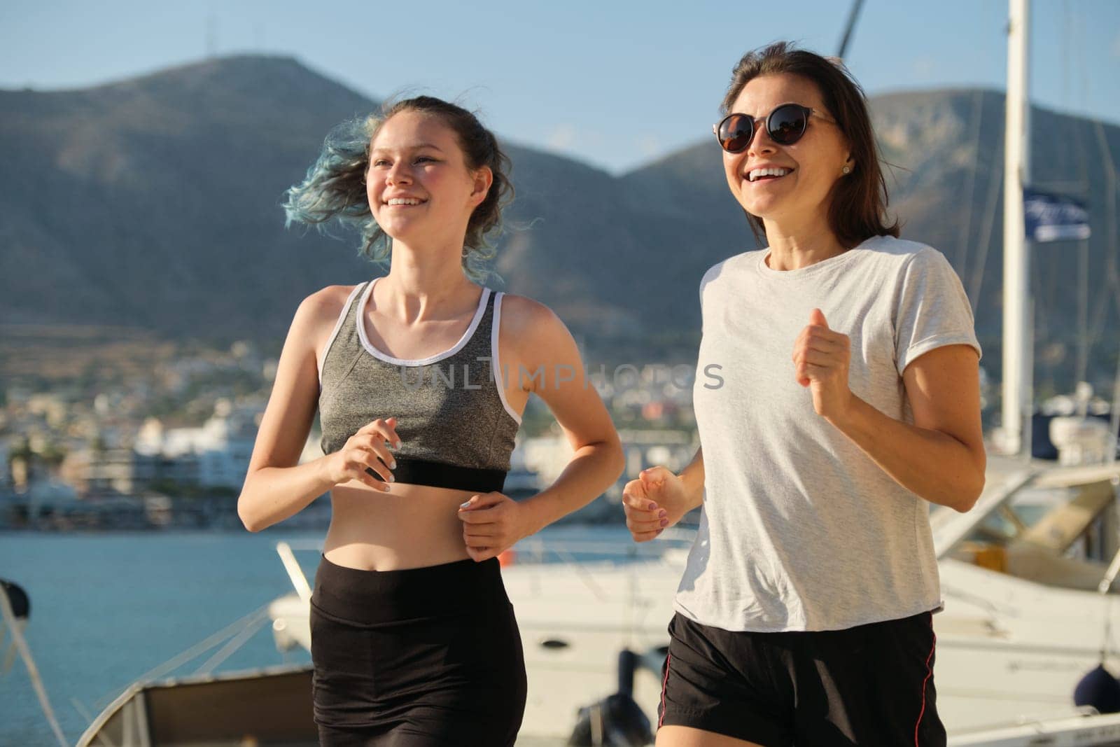 Smiling mother and daughter teenager jogging running at seaside promenade together, sports outdoor, background sea mountains city, sunny summer day