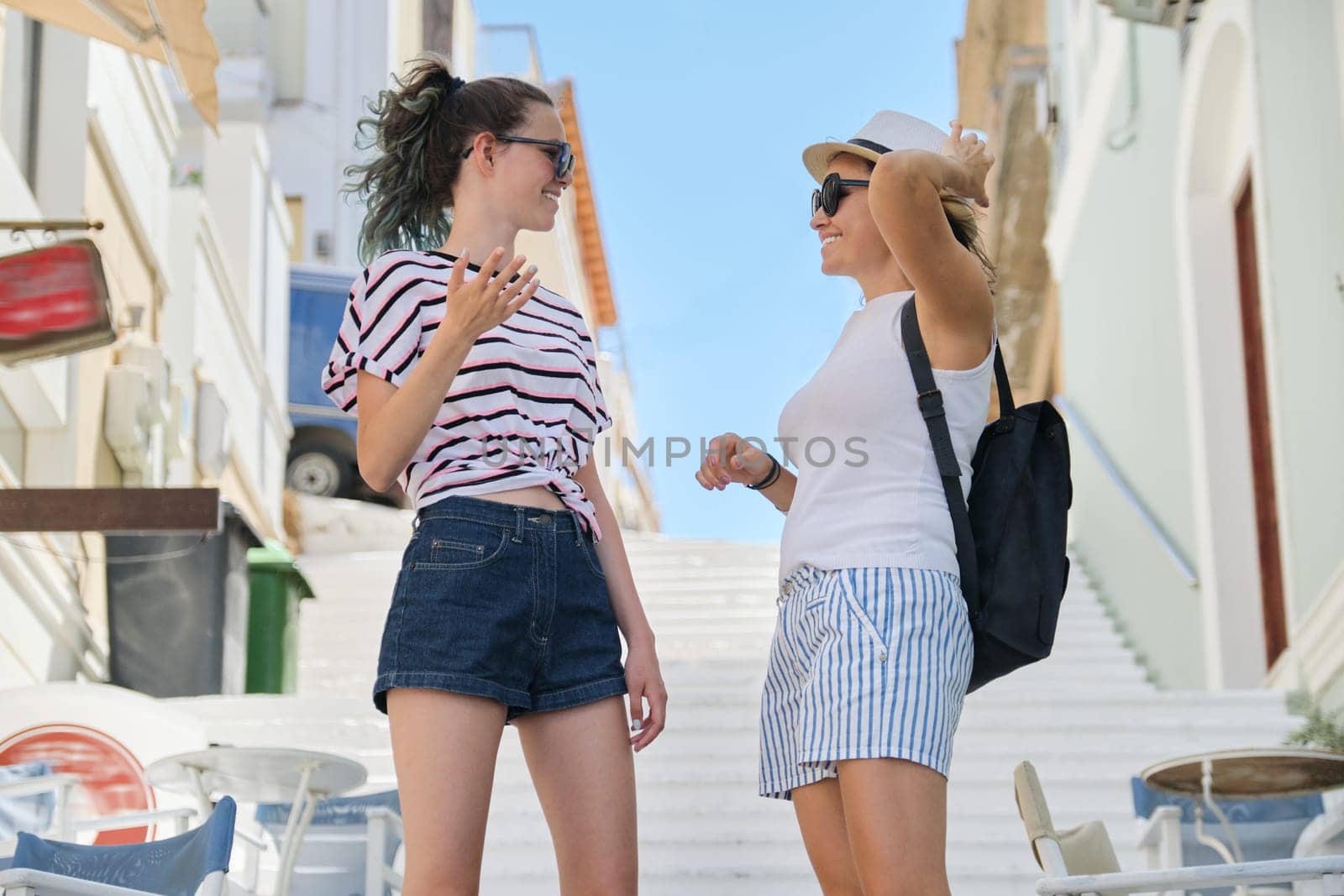 Portrait of mother and teenage daughter talking and smiling, summer city resort background white staircase