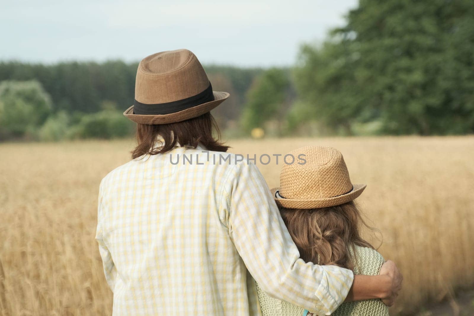 Mother hugging her little daughter back view, yellow ripe wheat field nature background, sunset golden hour