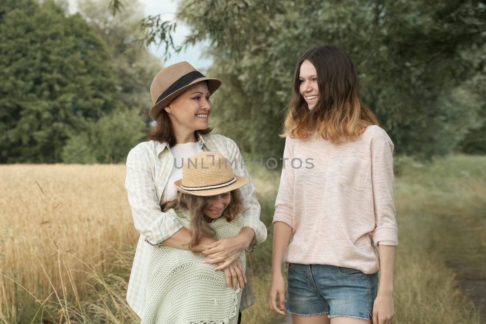 Happy loving family mother and children two daughters walking together along country road, summer nature background, wheat field, golden hour