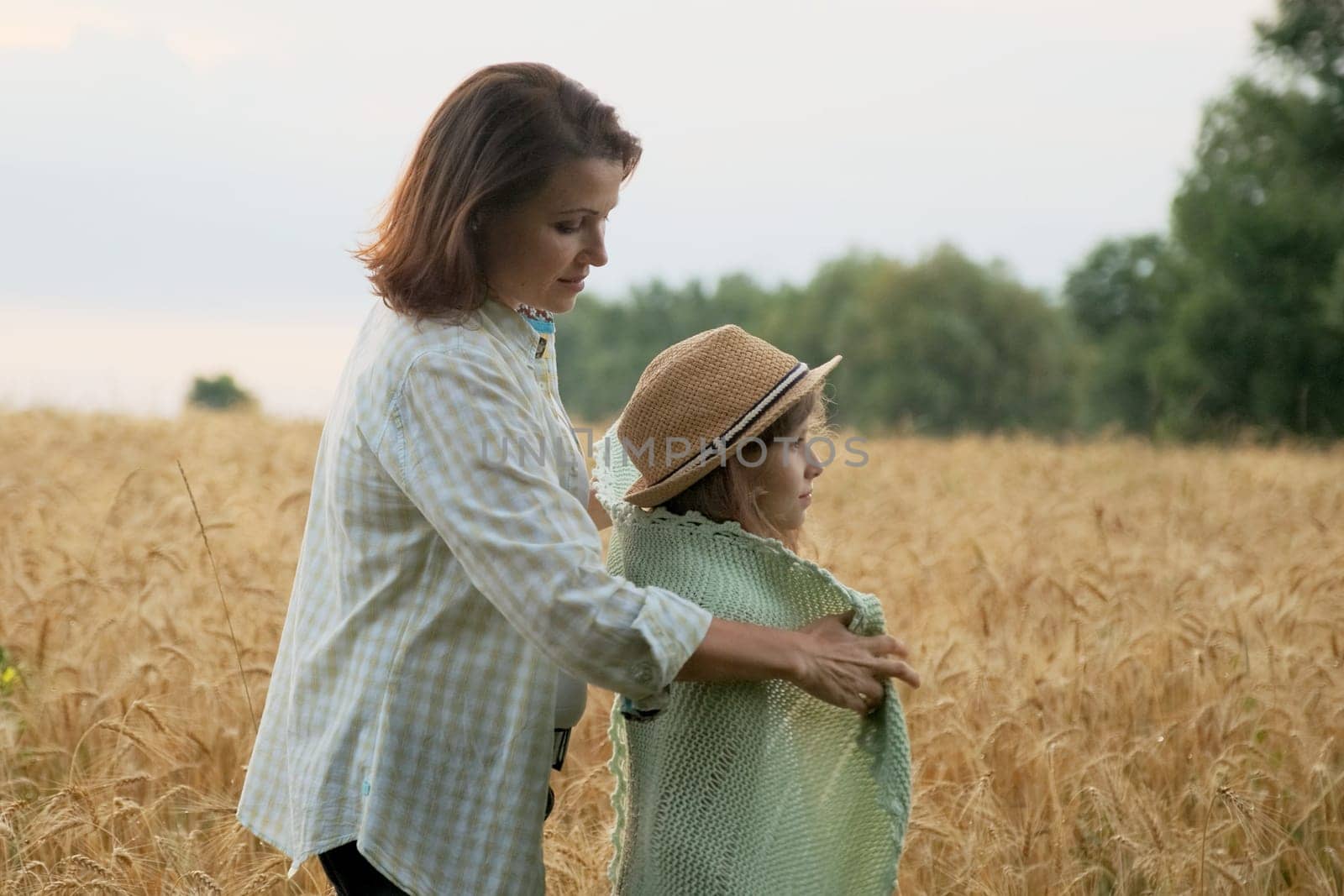 Loving moms puts on daughter knitted blanket, hugging child, yellow ripe wheat field nature background, golden hour