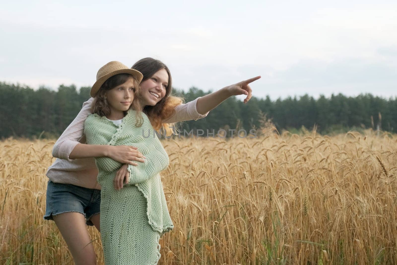 Elder sister hugged younger and shows a hand to the side, background field of yellow ripe wheat