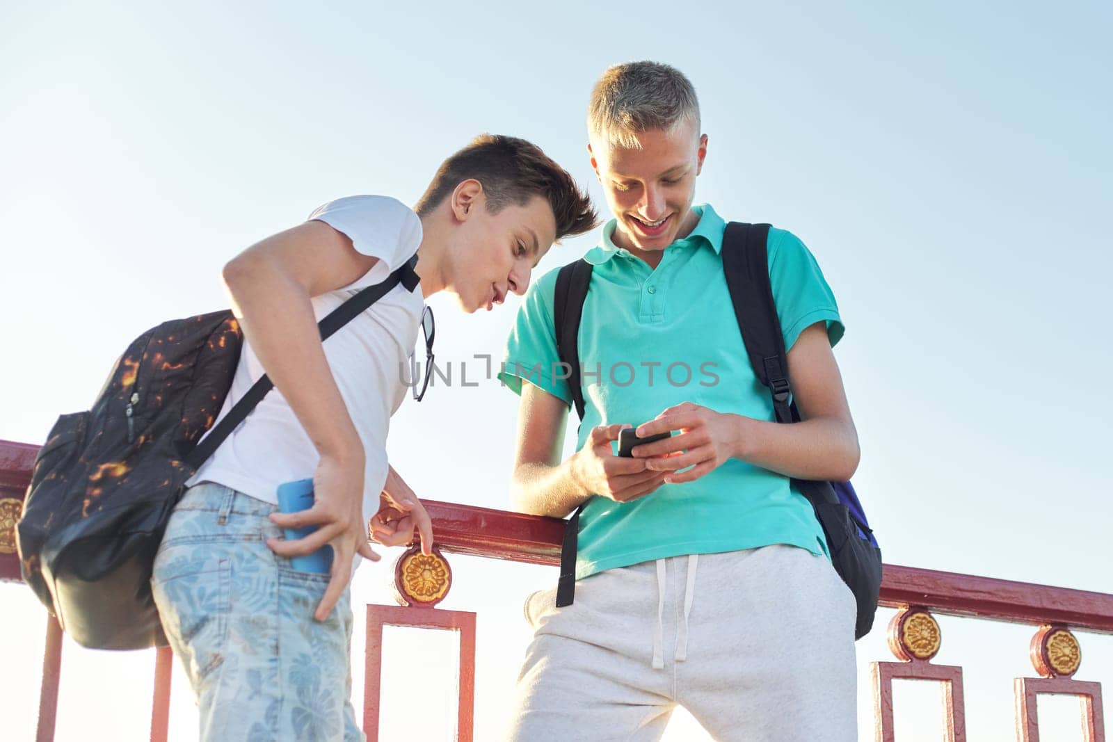 Outdoor portrait of two talking boys teenagers 15, 16 years old, males laughing look at smartphone, golden hour