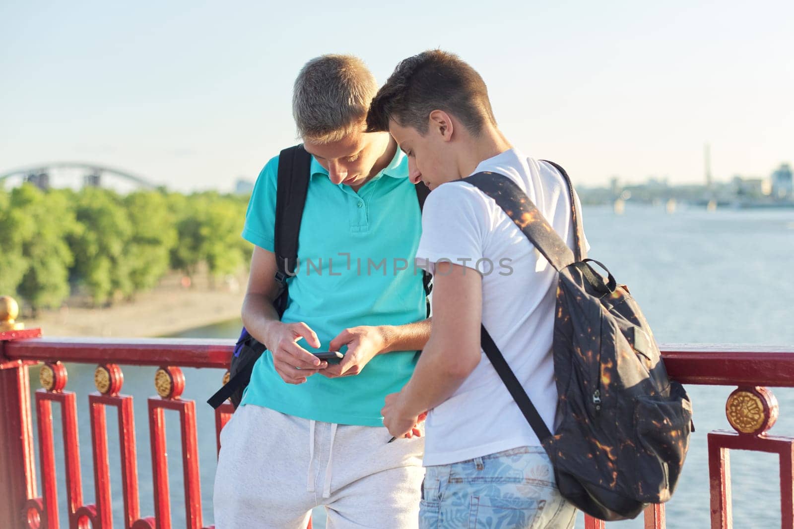 Two friends teenage boys using smartphone, talking and smiling, outdoor summer day, sunset on the bridge