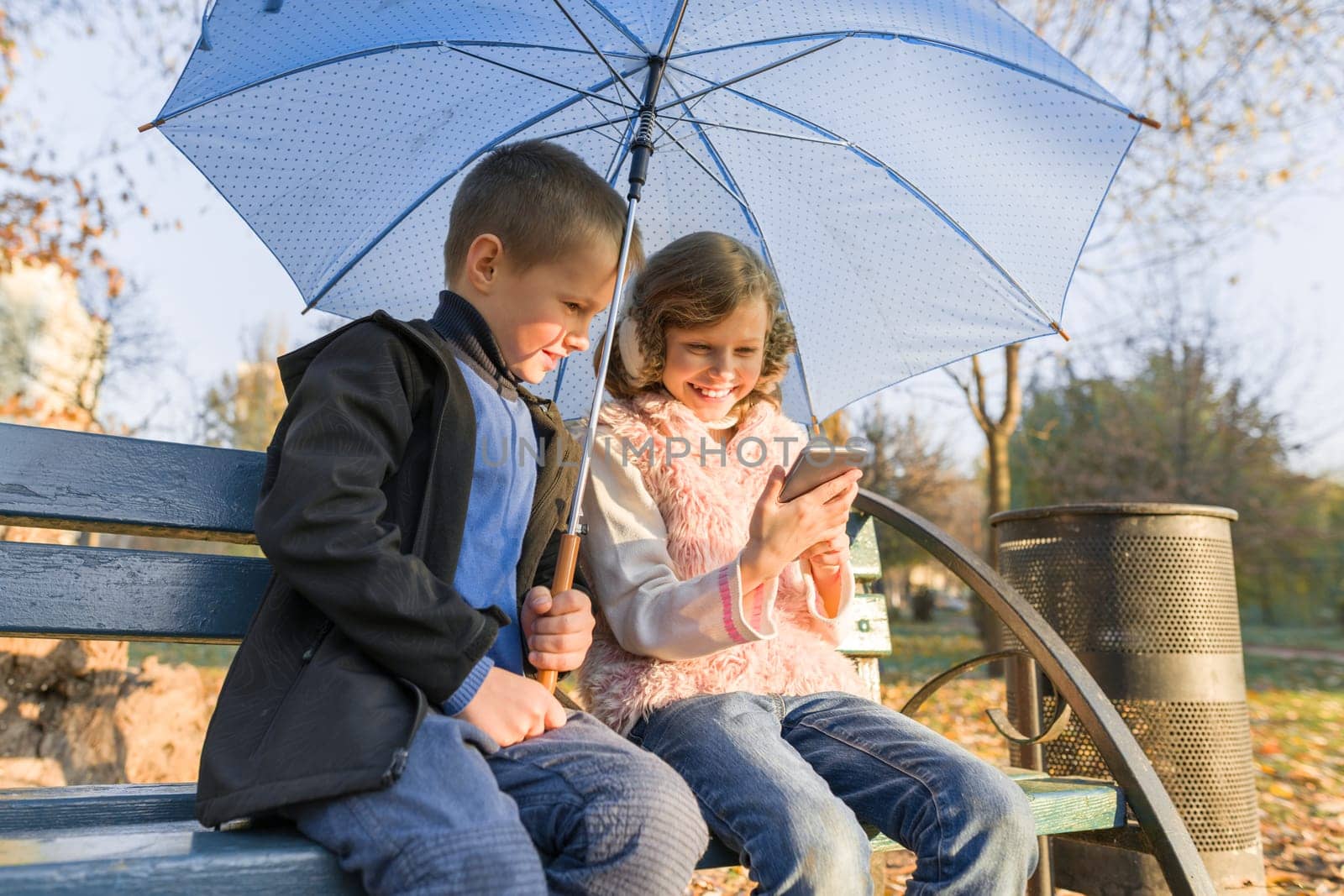 Outdoor portrait of two smiling children boy and girl, sitting under an umbrella on bench in autumn park, using smartphone, golden hour