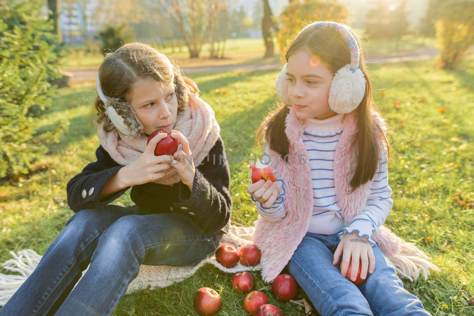 Children two girls eating red apples sitting in yellow autumn park, golden hour