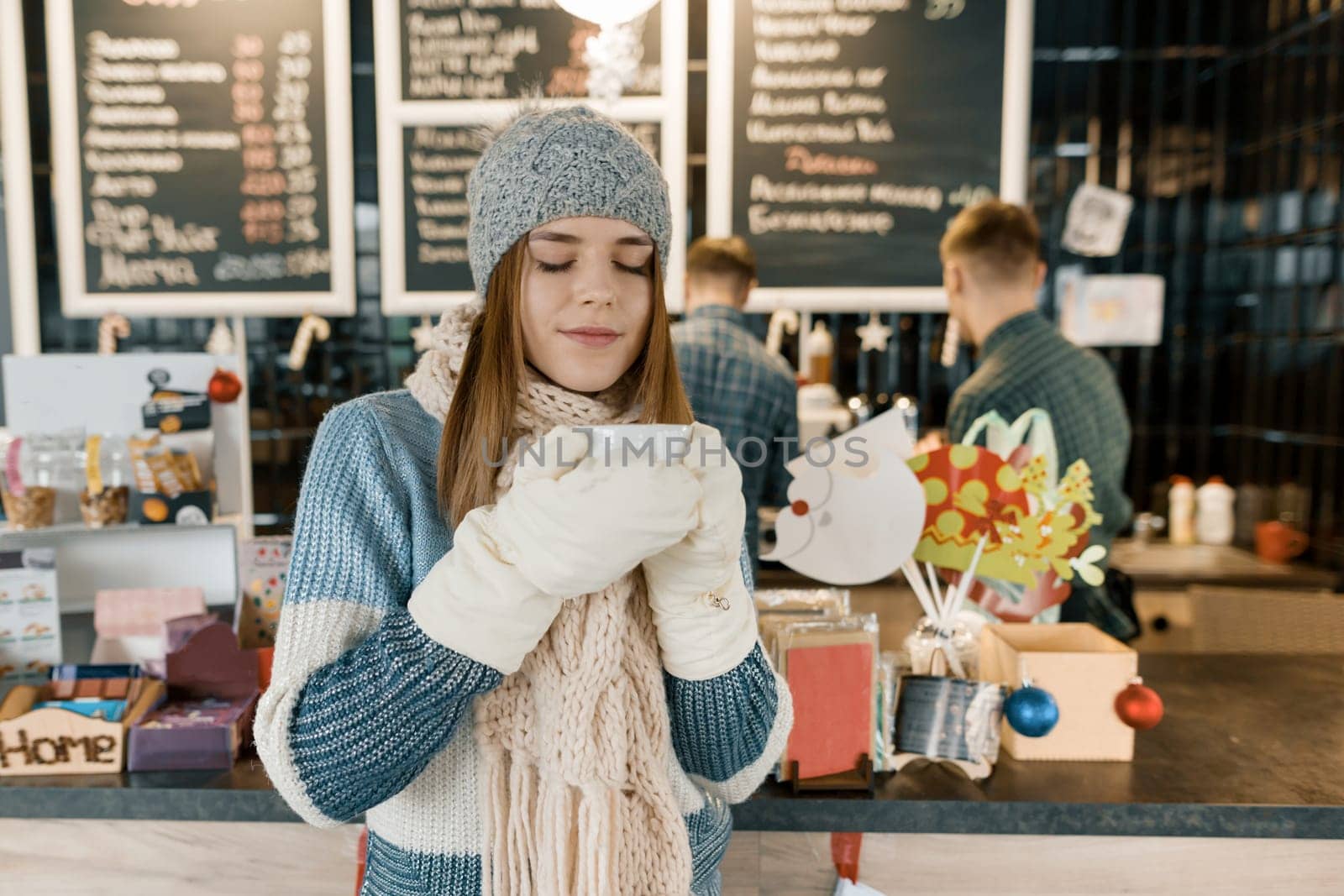 Winter portrait of young beautiful woman in knitted scarf, knitted hat, mittens, warm sweater with cup of coffee. Girl near bar counter, Christmas decorated tree in coffee shop.