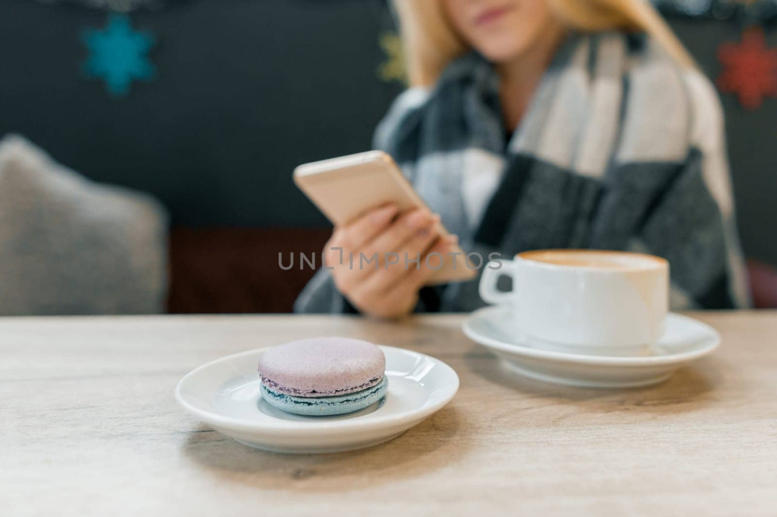 Macaroon in plate, cup of coffee, beautiful blonde girl in woolen blanket with smartphone in the interior of winter coffee house, focus on macaroons.