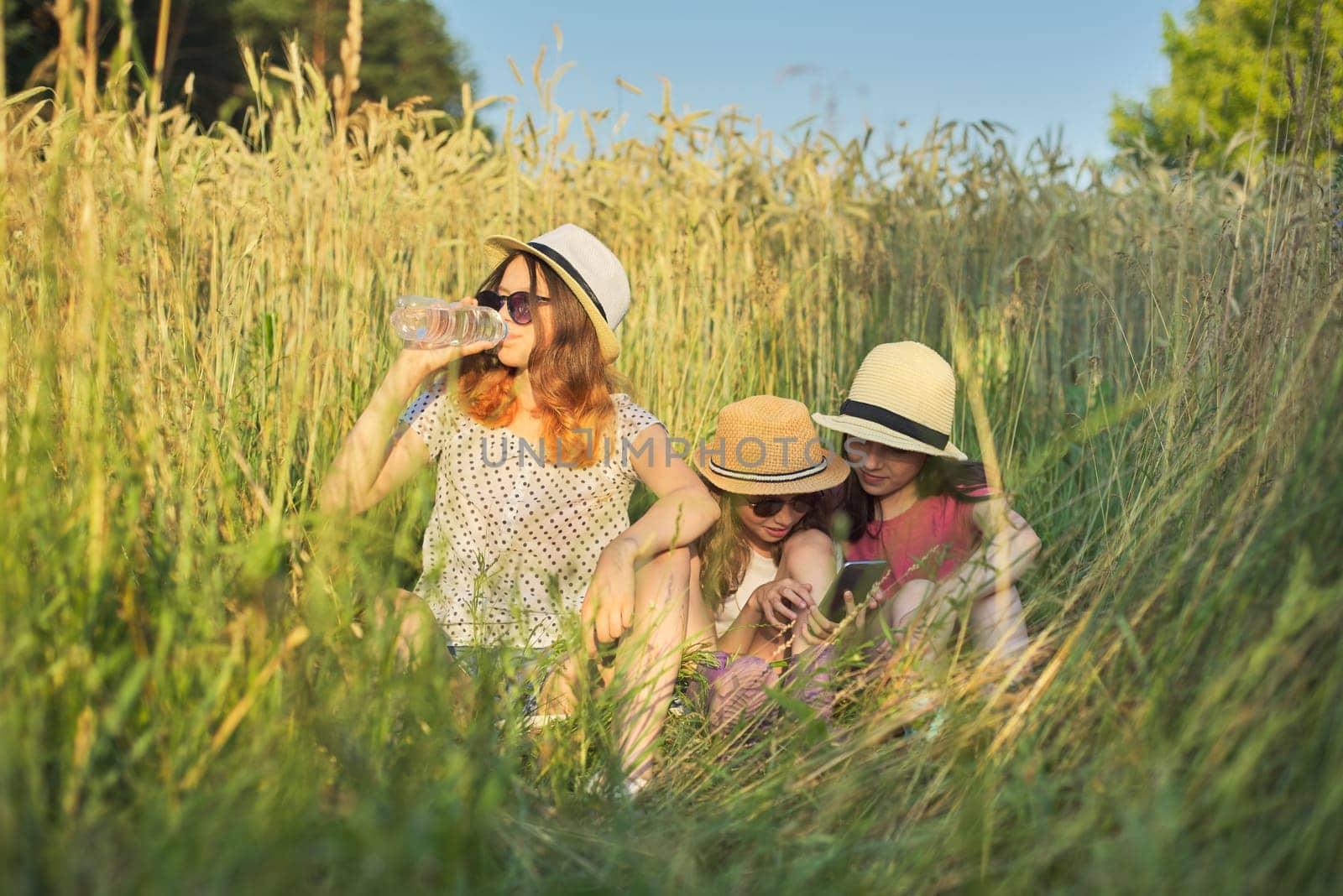 Portrait of three girls sitting and resting in grass, children talking, looking at smartphone laughing on sunny summer day, sunset in meadow