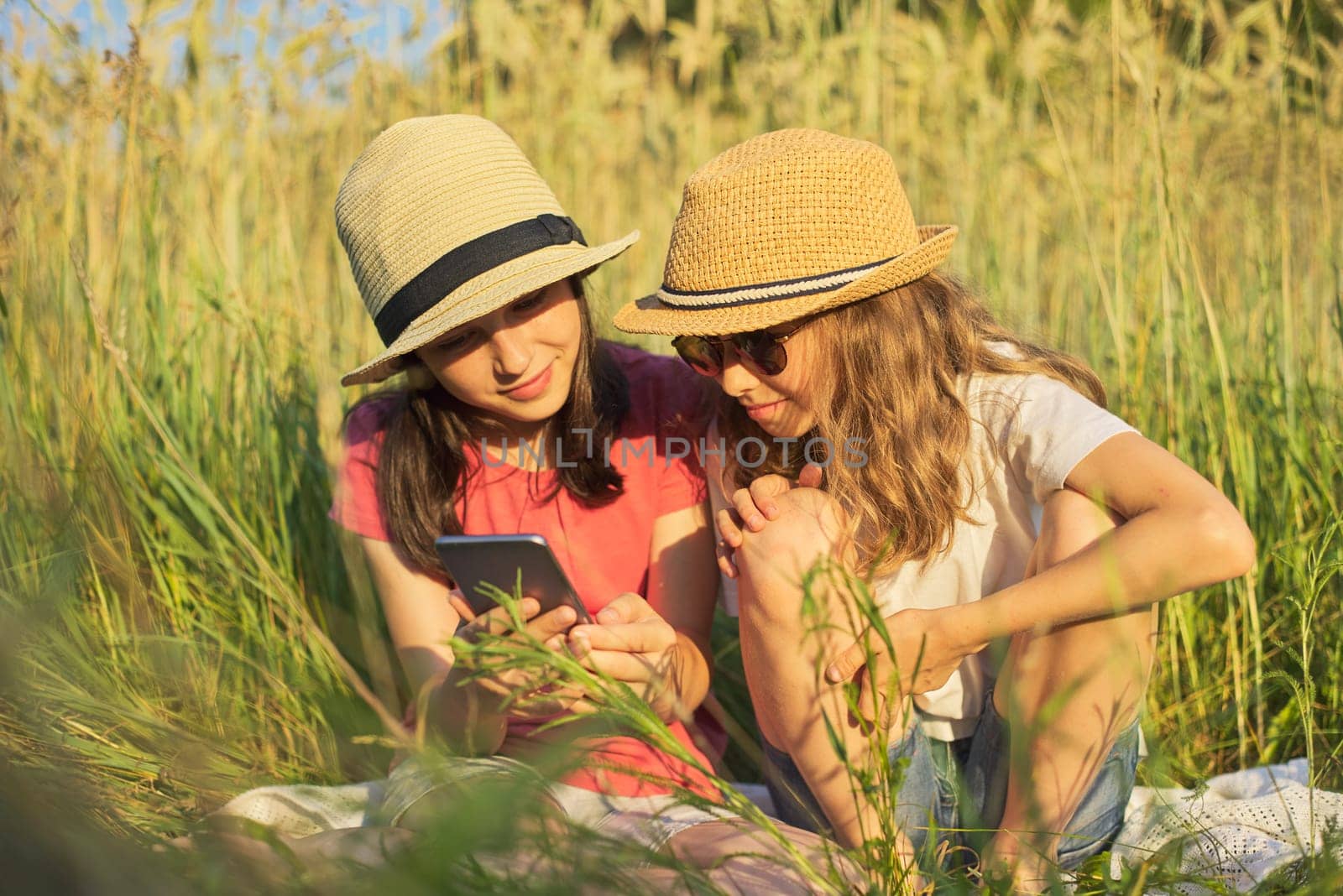 Summer portrait of two girls sitting in grass playing and looking at smartphone, beautiful children at sunset