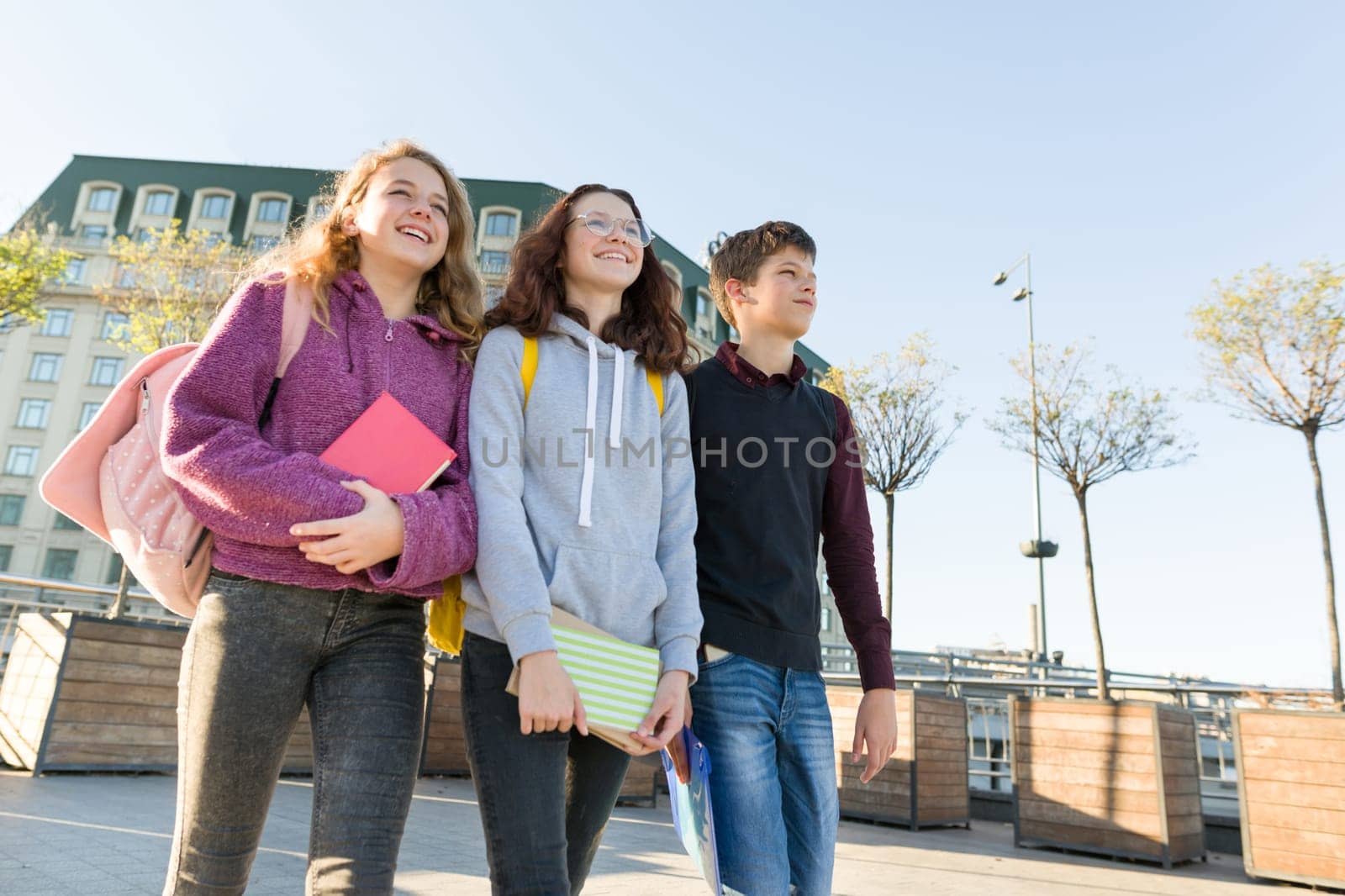 Smiling teens students with backpacks and textbooks, talking and going forward. City background, golden hour