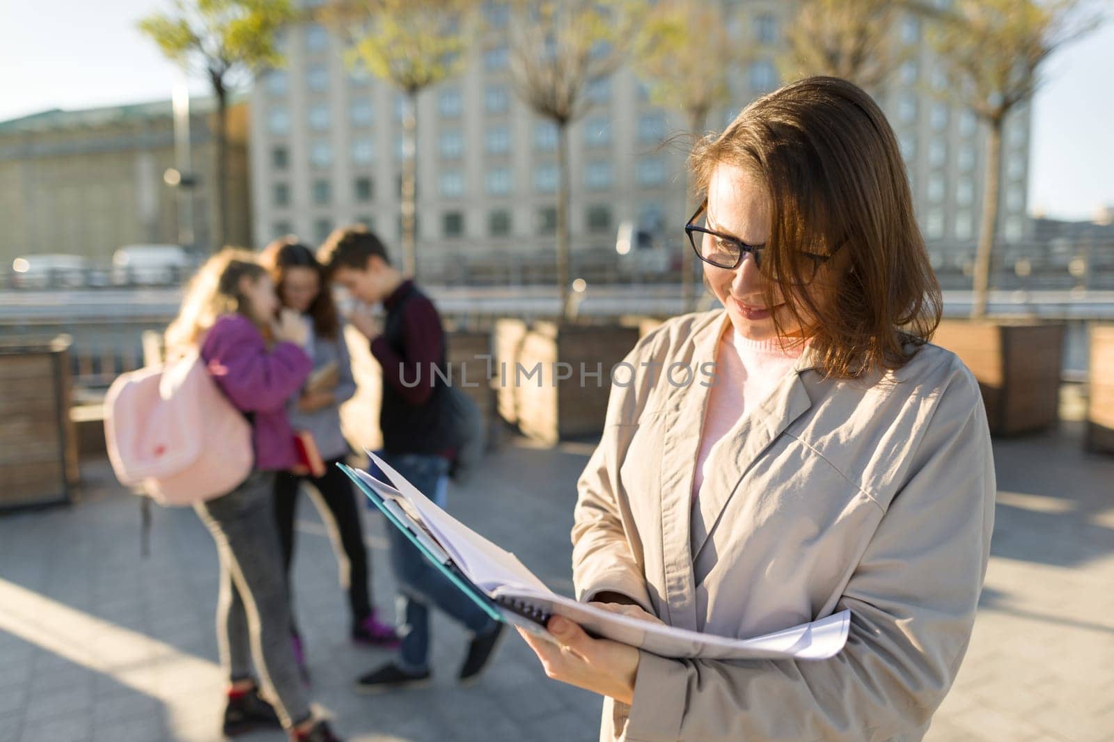 Portrait of mature smiling female teacher in glasses with clipboard, outdoor with a group of teenagers students, golden hour