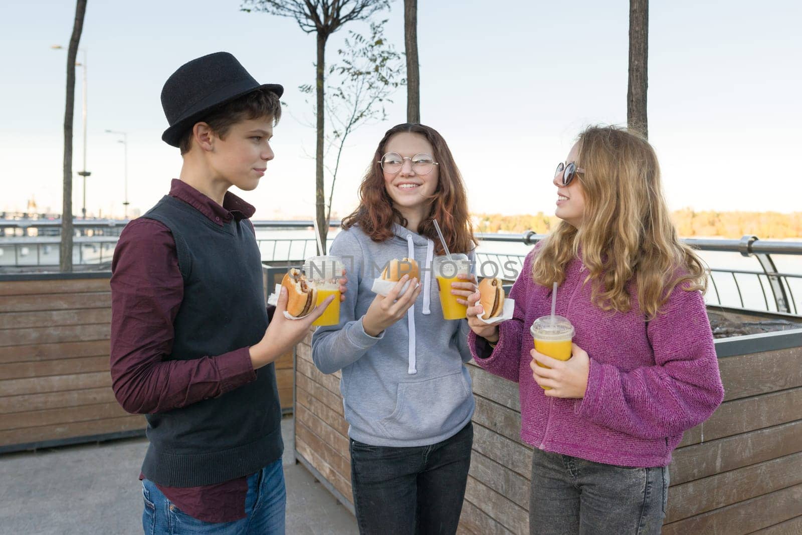 Teenagers eat street food, friends boy and two girls on city street with burgers and orange juice. City background, golden hour