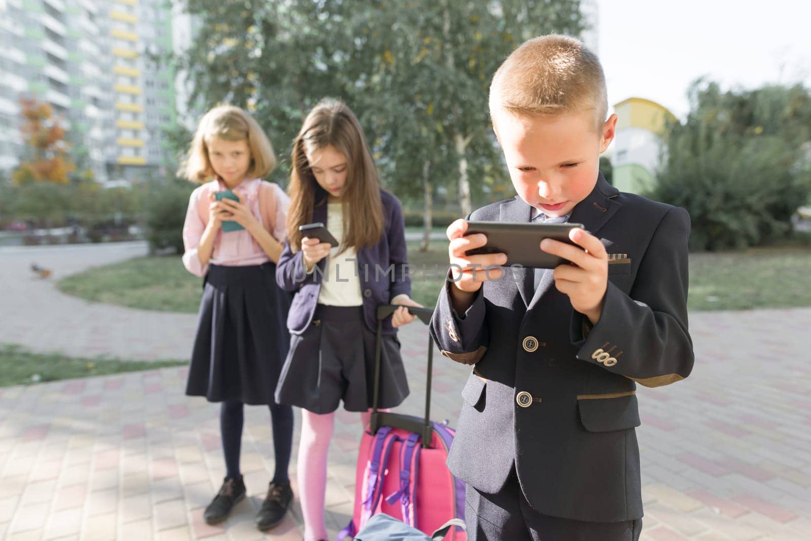 Children of elementary age with smartphones, backpacks, outdoor background. Education, friendship, technology and people concept