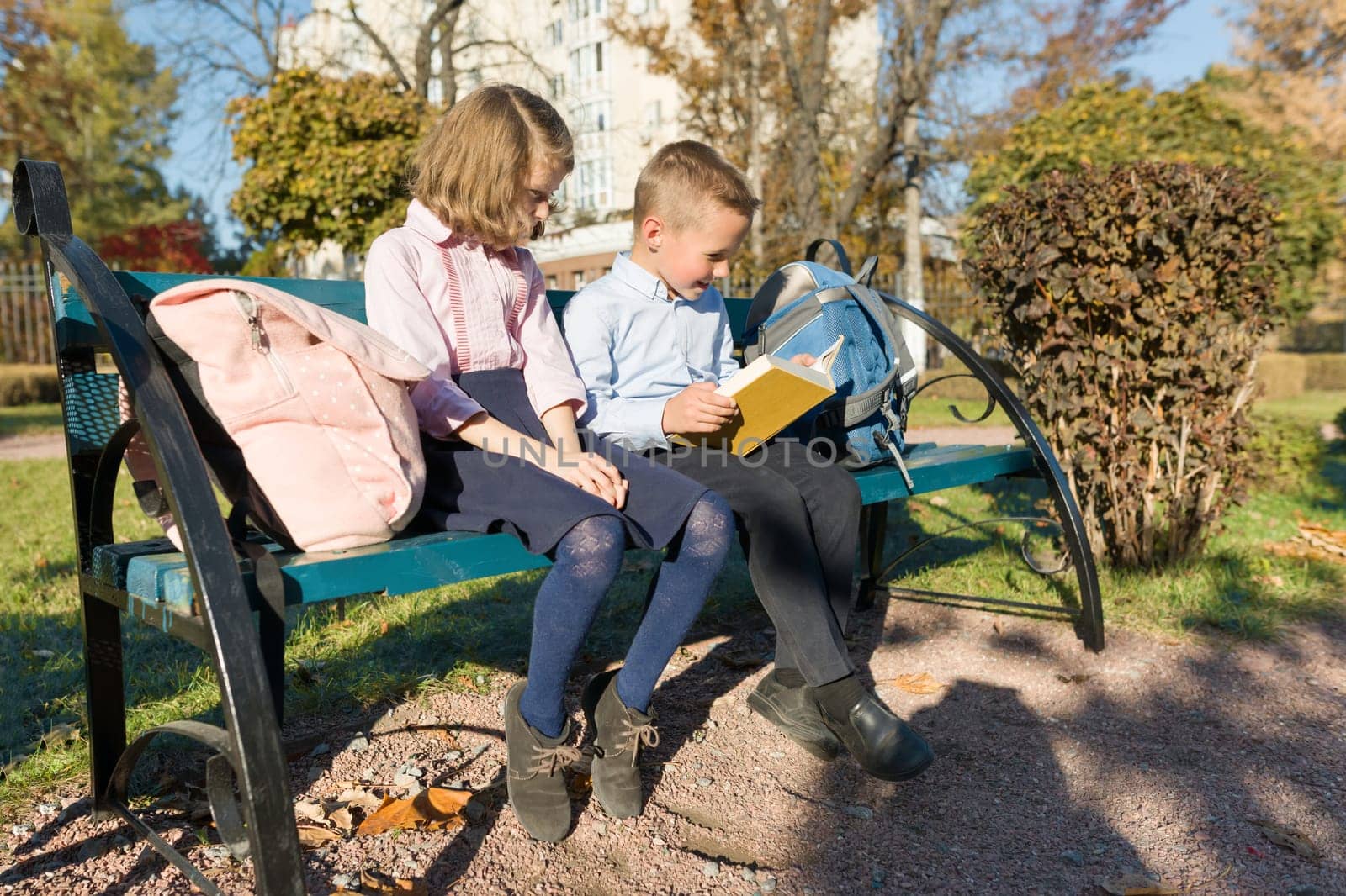 Little boy and girl schoolchildren reading book, sitting on bench, children with backpacks, bright sunny autumn day