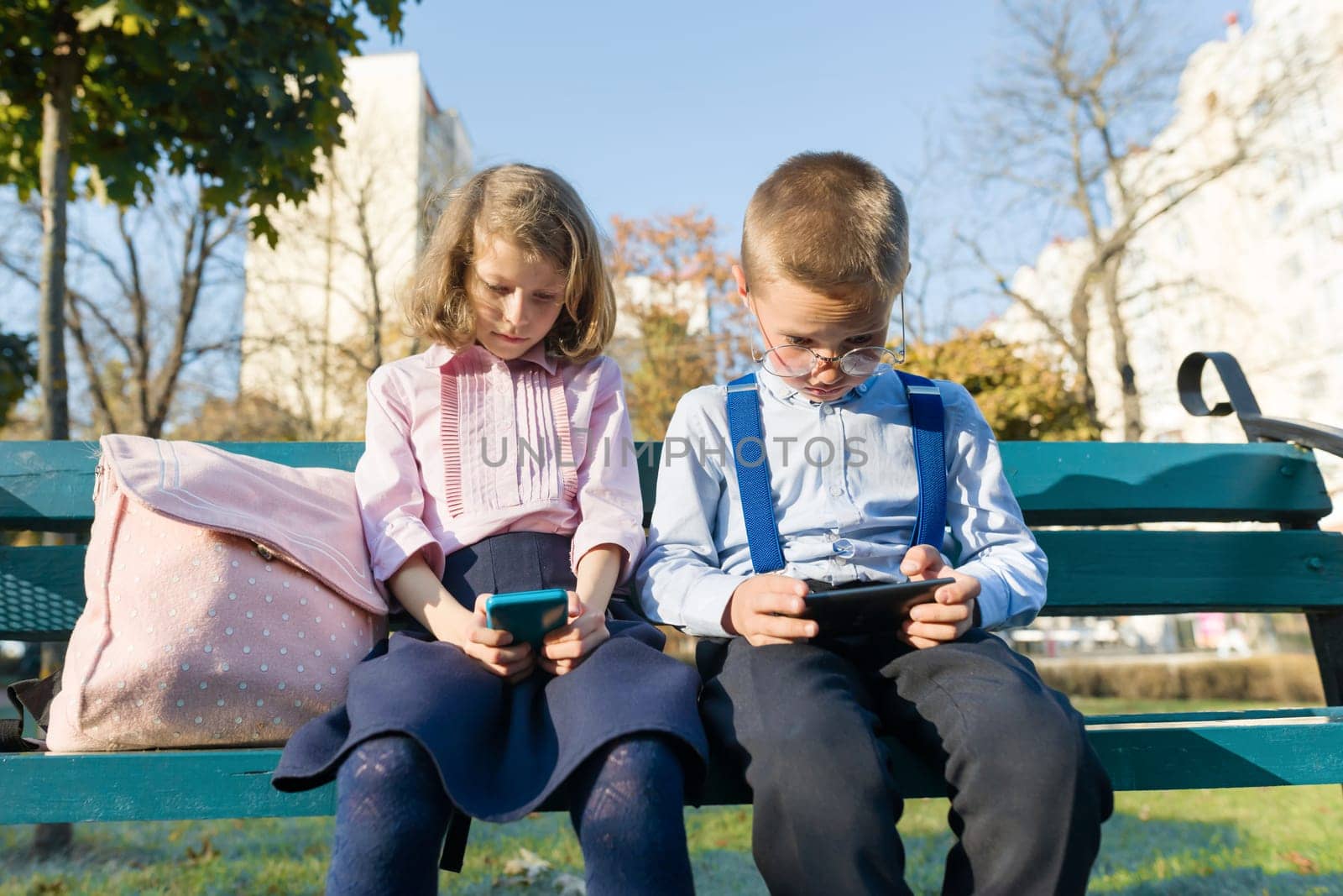 Smart serious children boy and girl are looking into smartphones. On a bench with school backpacks, background autumn sunny park, golden hour