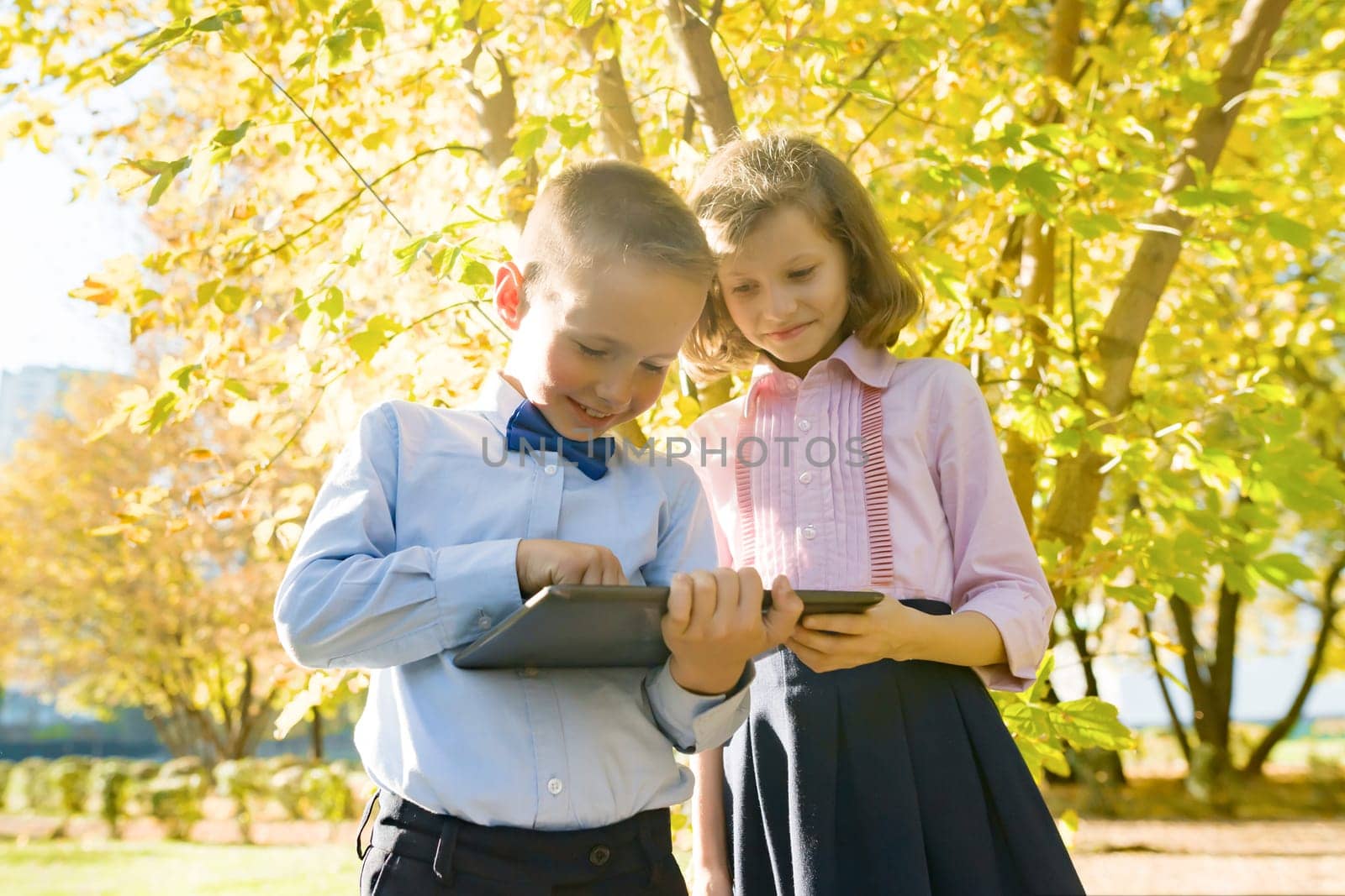 Two kids watching digital tablet, background autumn sunny park, golden hour