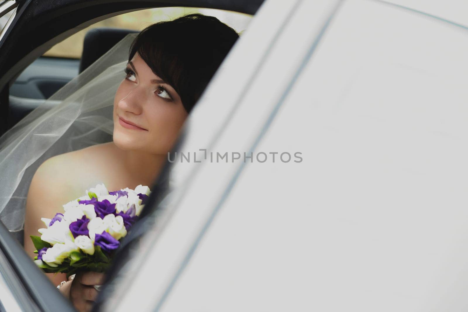 A young brunette bride is sitting in a car in close-up with a colorful bouquet of flowers on a summer day